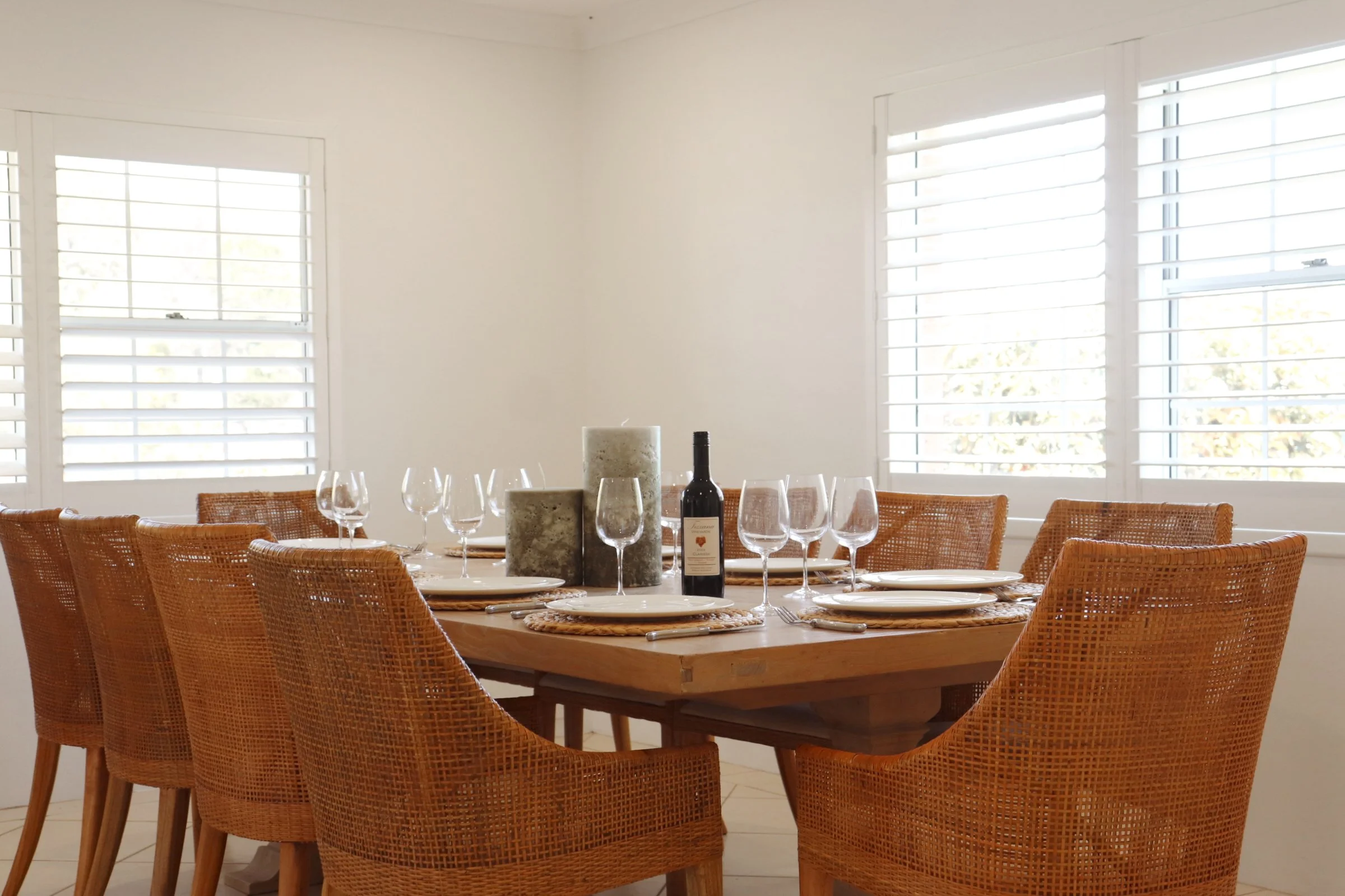 A dining table set for a meal with wine glasses, plates, and candles in a bright room with white walls and window shutters.