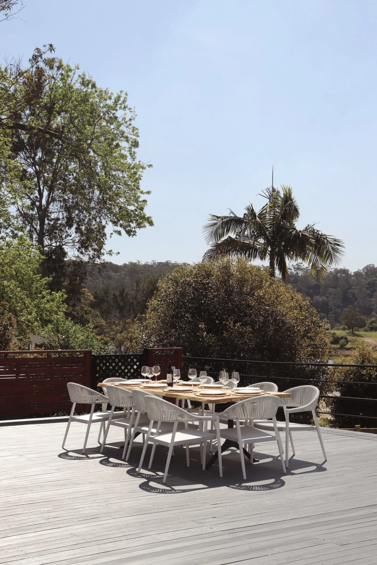 An outdoor dining table set with plates, wine glasses, and a wine bottle on a wooden deck, surrounded by trees and under a clear blue sky.