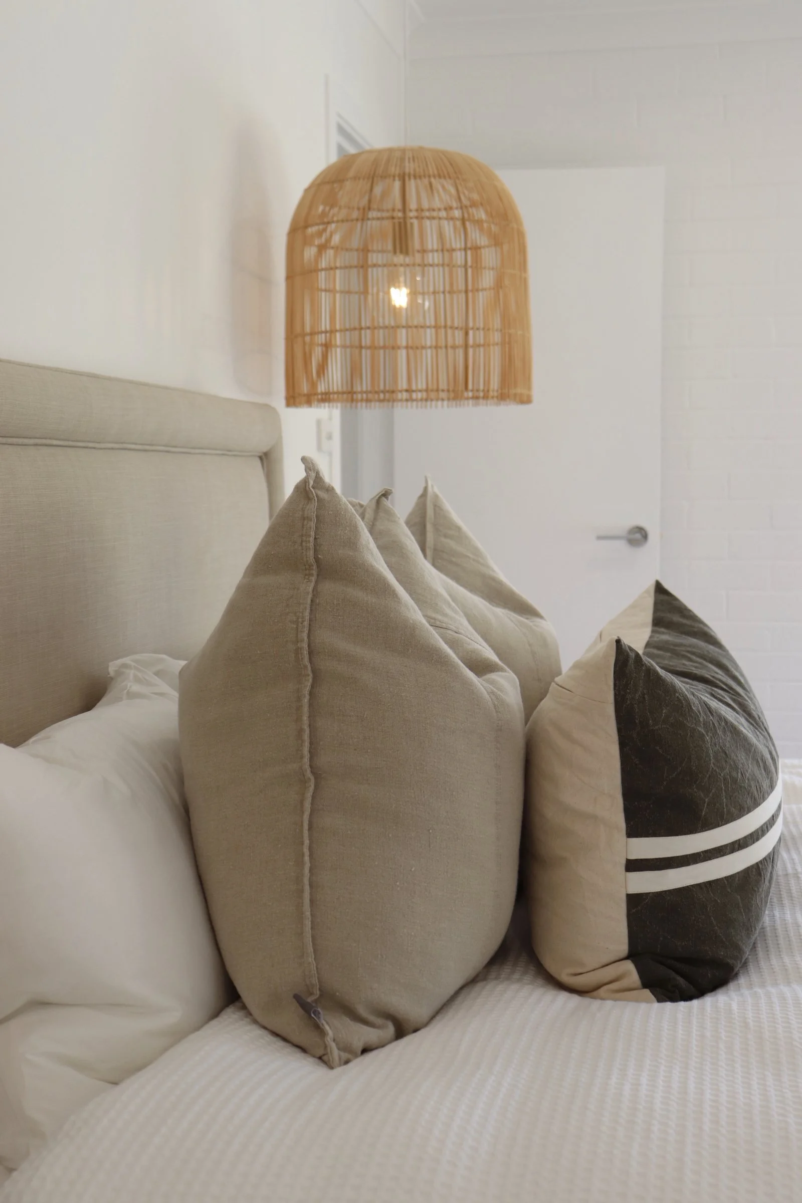 Close-up of a bed with beige, black, and white decorative pillows, a beige headboard, and a woven rattan pendant light hanging above.