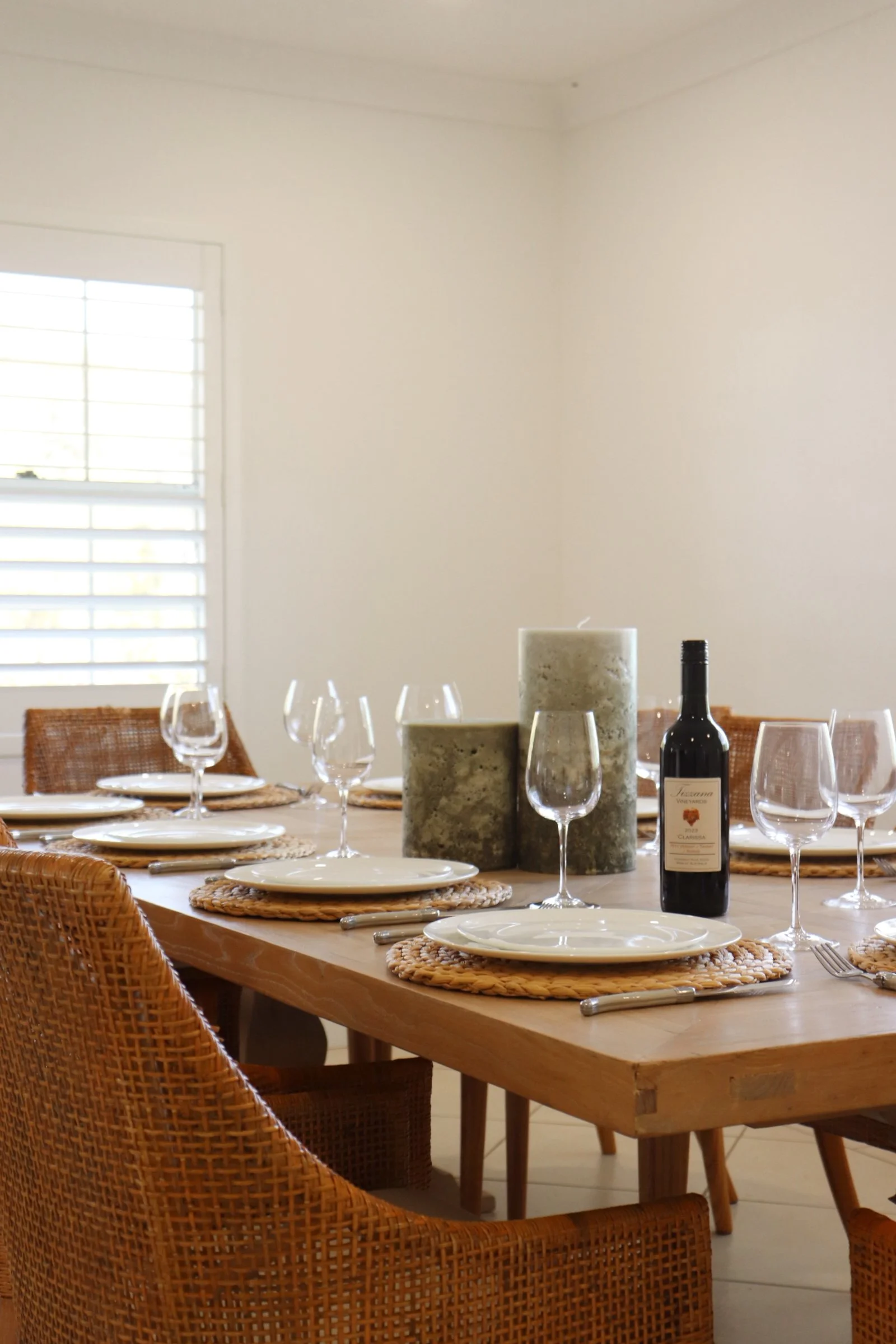 A dining table set for a meal, with white plates on woven placemats, wine glasses, cutlery, two large candles, and a bottle of red wine in a bright room with a window and white walls.