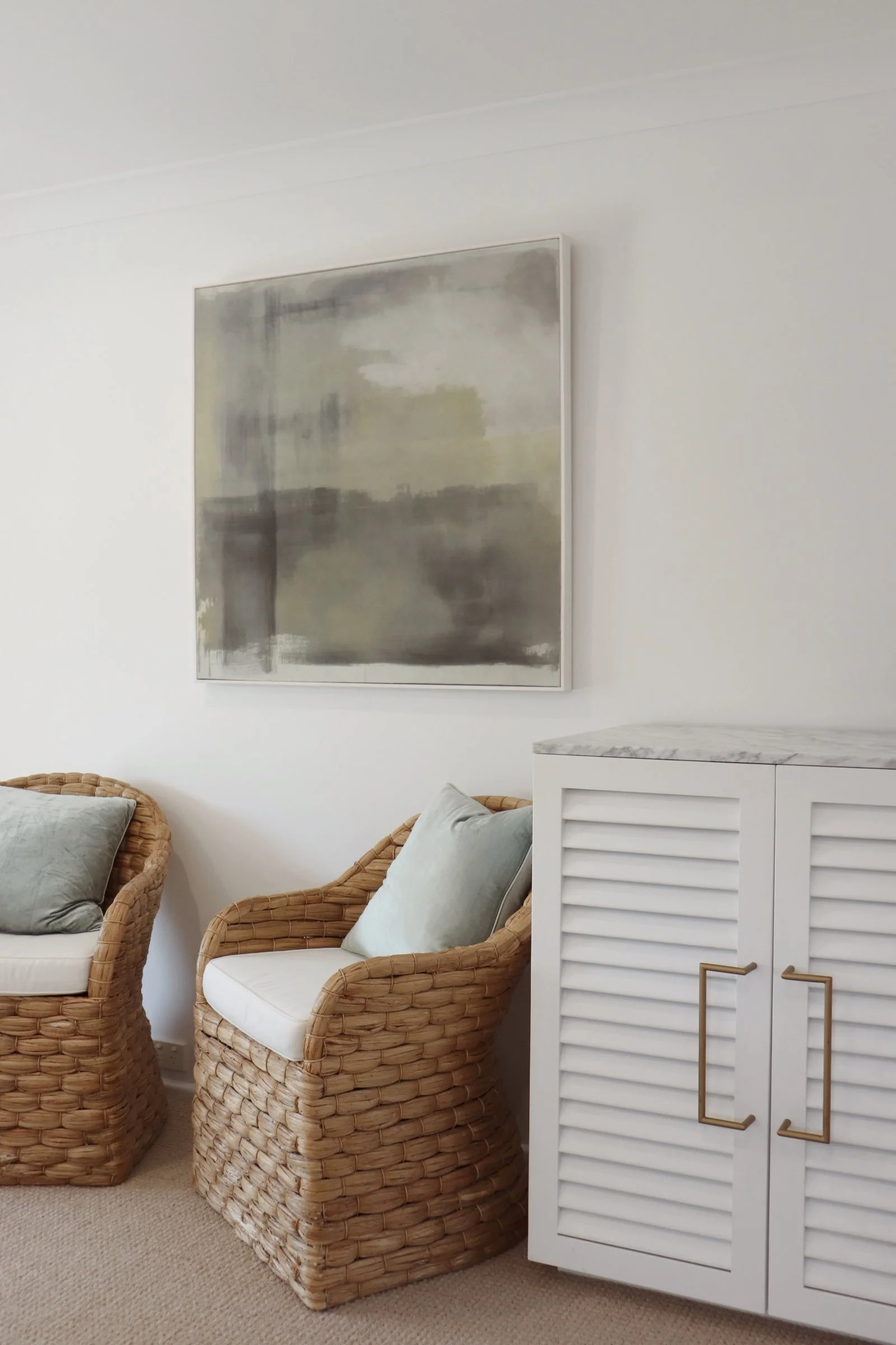 Living room corner with two rattan armchairs with cushions, white cabinet with marble top, and abstract painting on white wall.