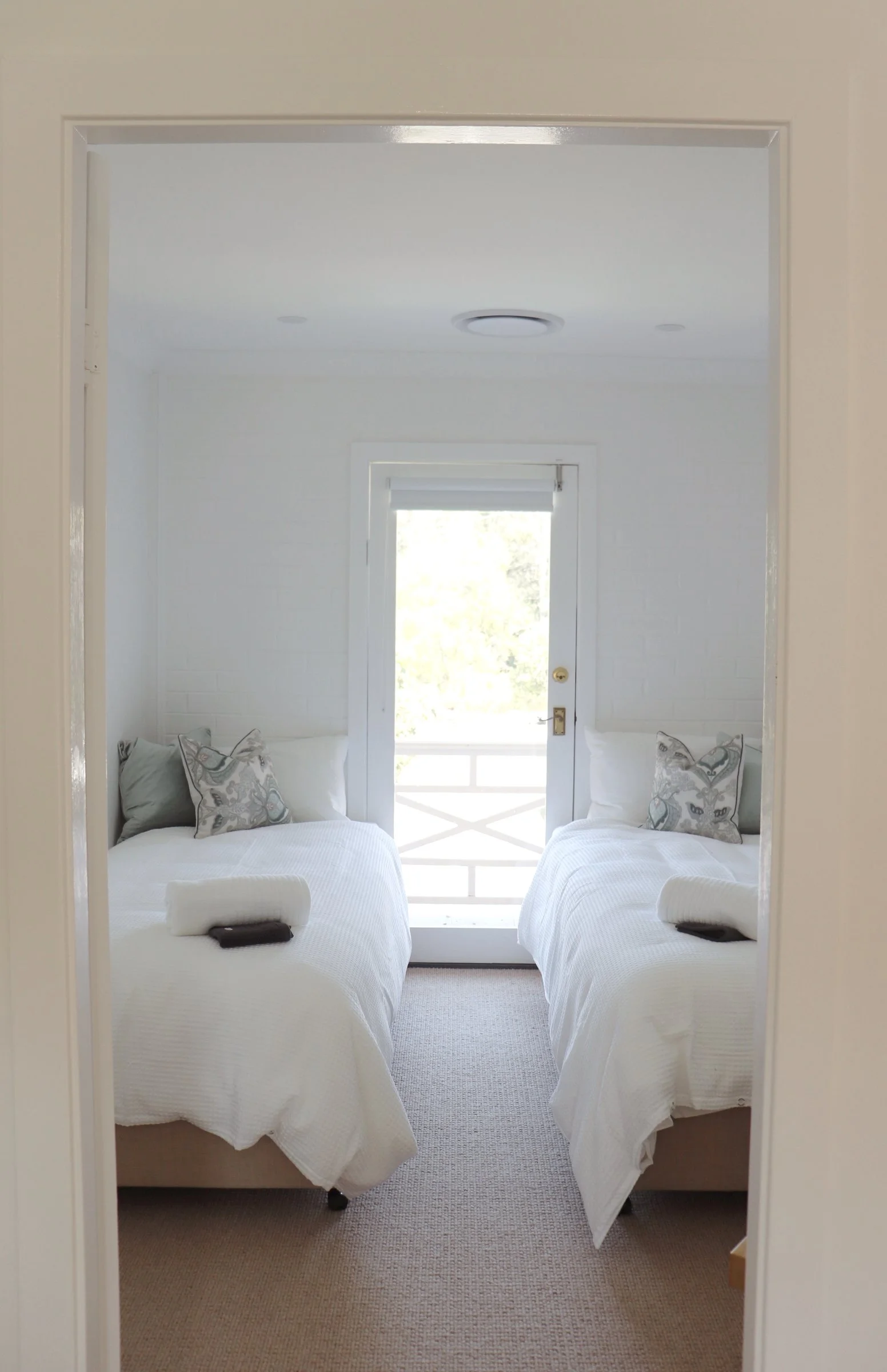 Room with two beds covered in white bedding, each with a rolled towel and a guest check-in card, small decorative pillow, and positioned on a beige carpet. A door at the far end leads to a balcony with a white railing, and a large window or door with