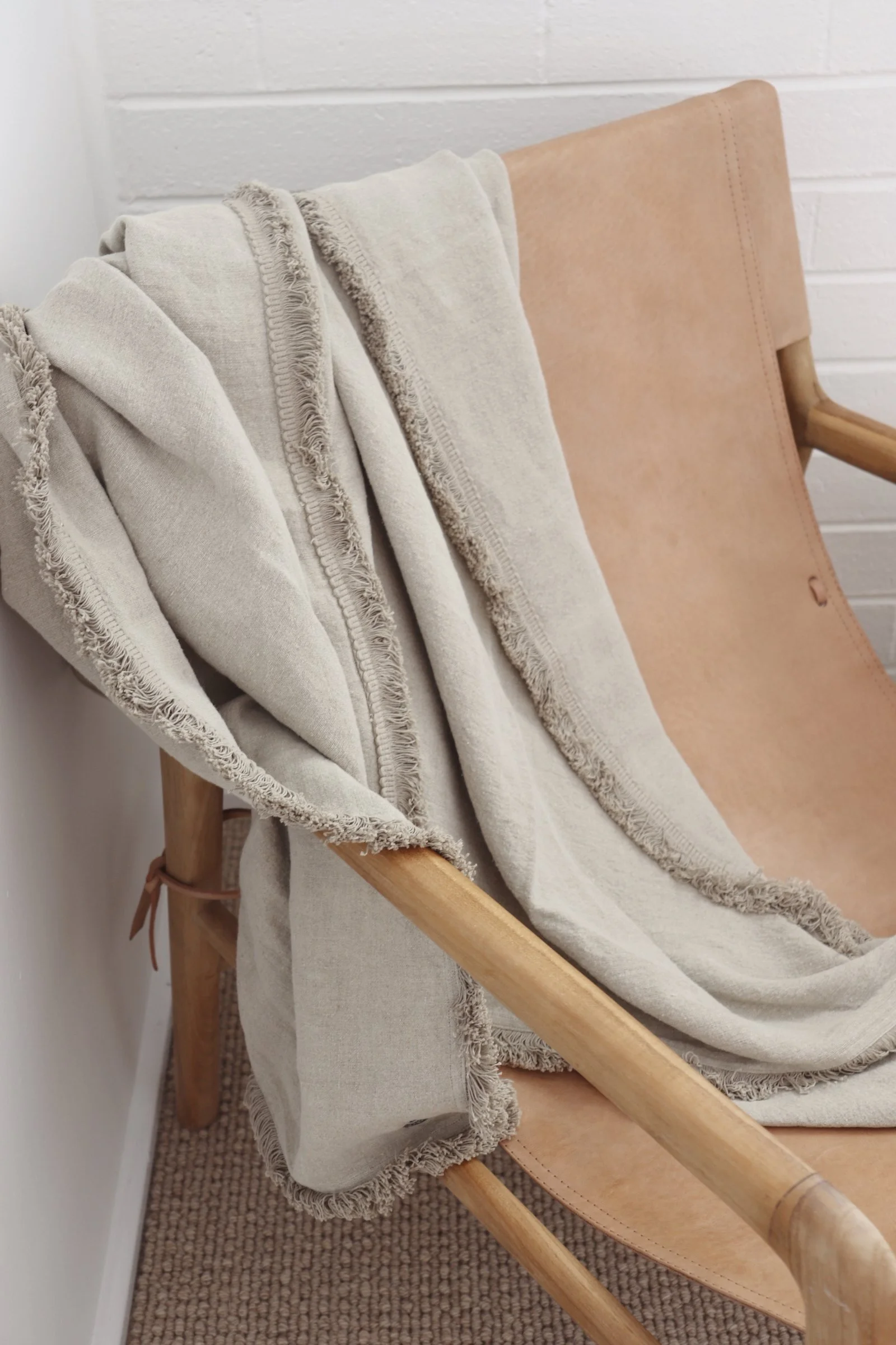A beige blanket with frayed edges draped over a light wooden chair with a leather seat, against a white brick wall.