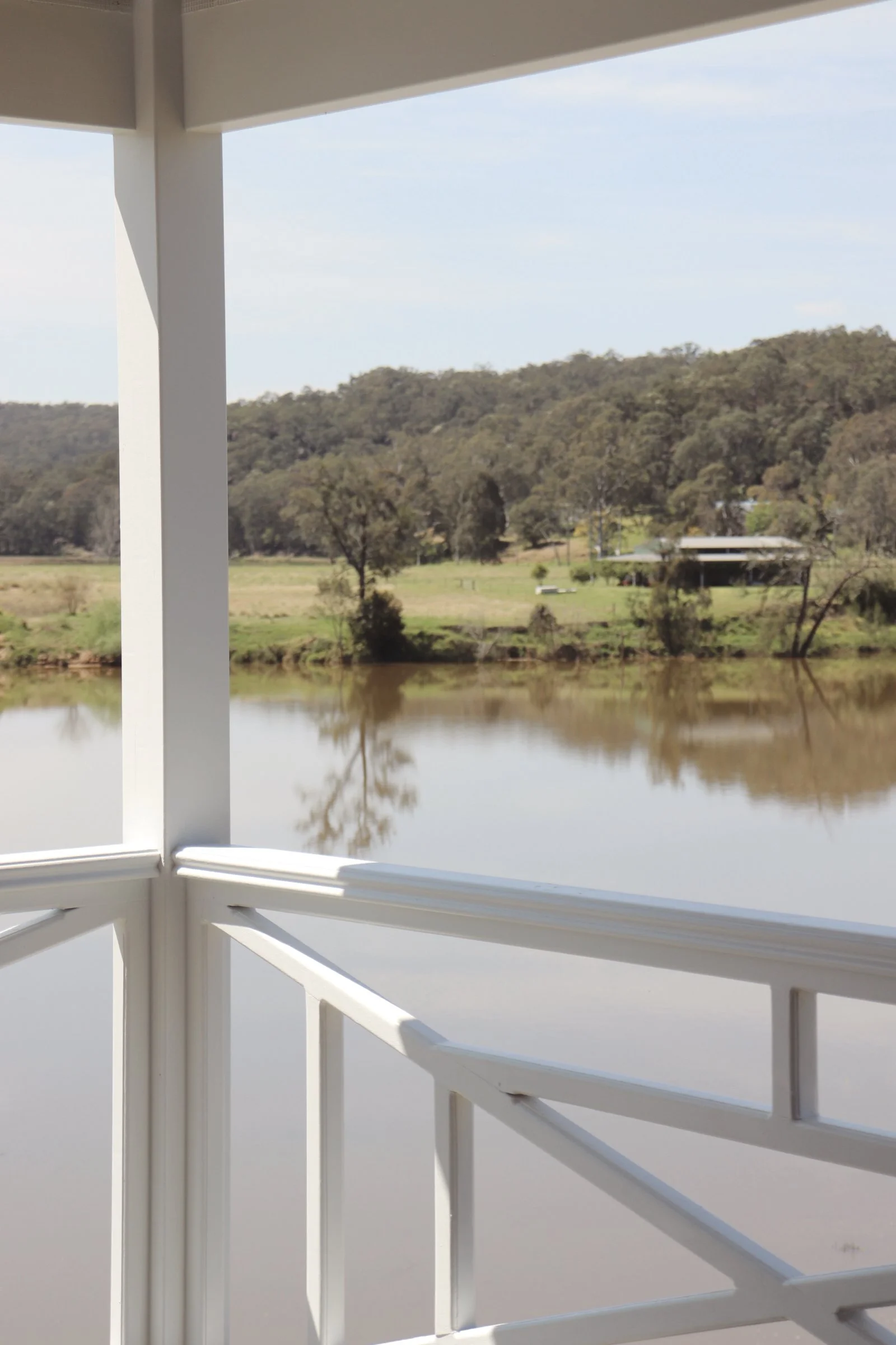 A view from a porch showing a calm river, trees, and a house on the far bank with hills in the background.