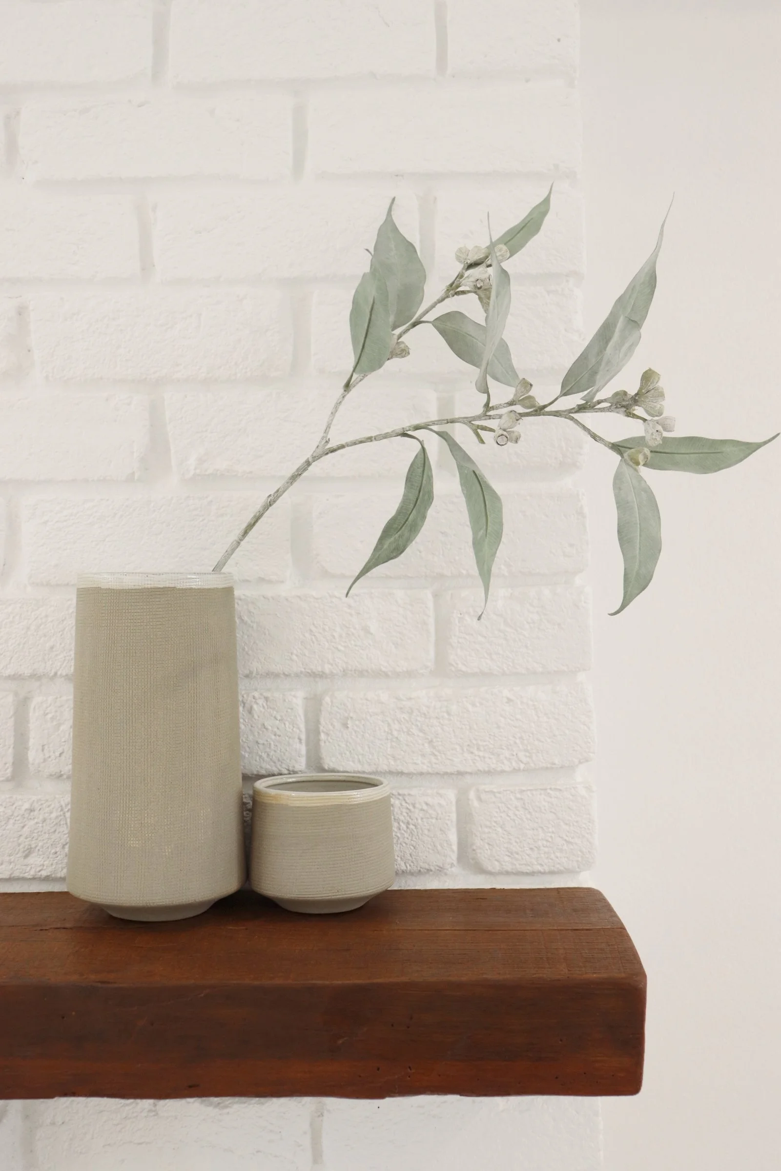 A beige textured vase with a long, slender neck holding a branch with large, pale green leaves and small buds, placed next to a smaller matching container on a wooden surface against a white brick wall.