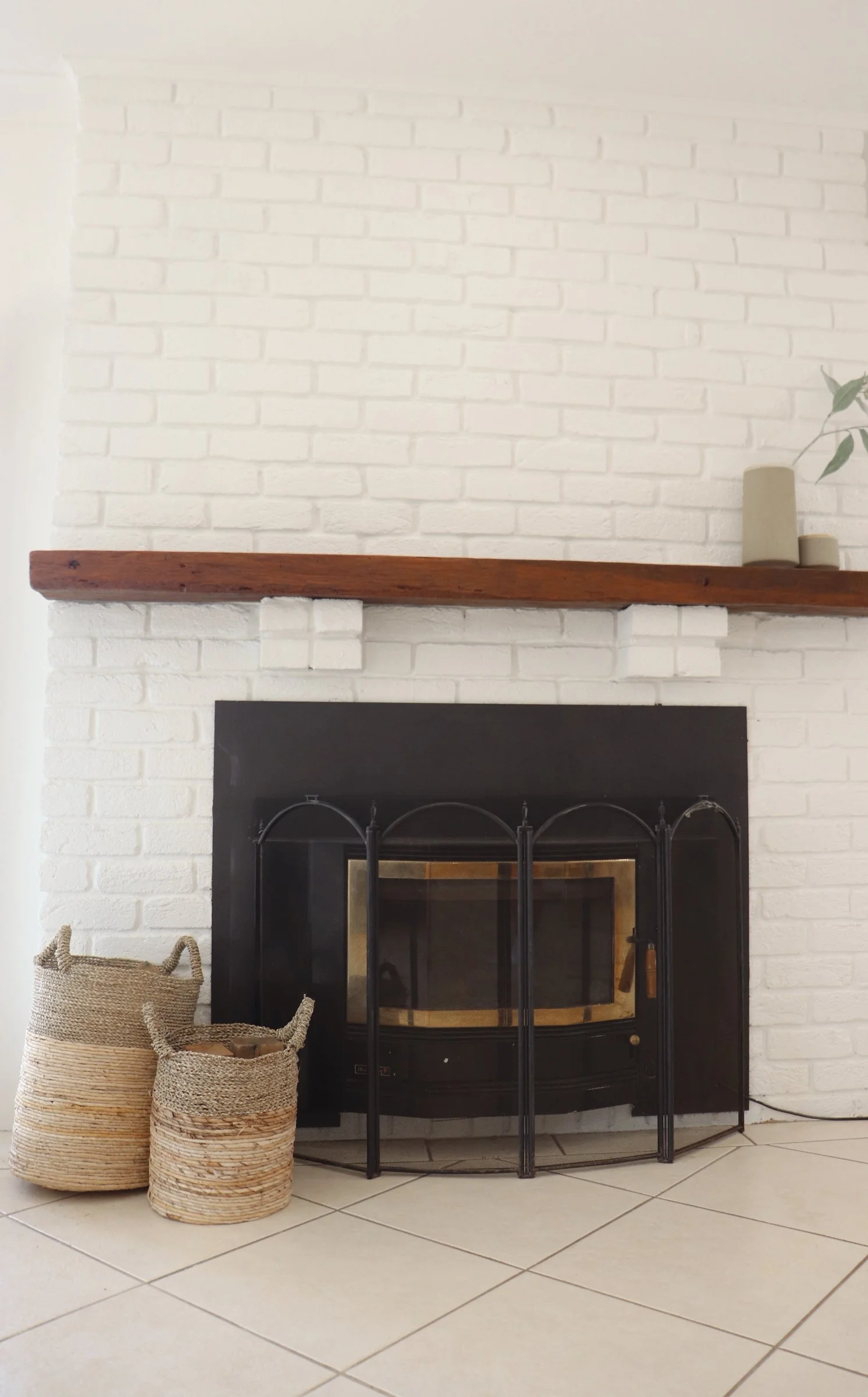White brick fireplace with black metal guard and wood mantel, two woven baskets on beige tiled floor, and minimal decor including three small vases on the shelf.