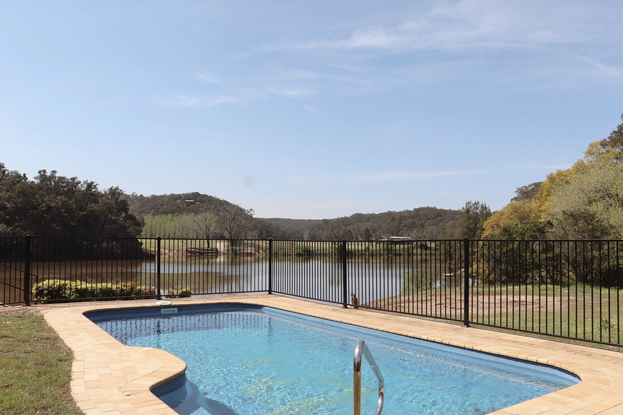 A backyard swimming pool with a black metal fence, overlooking a calm lake and hilly landscape under a partly cloudy sky.