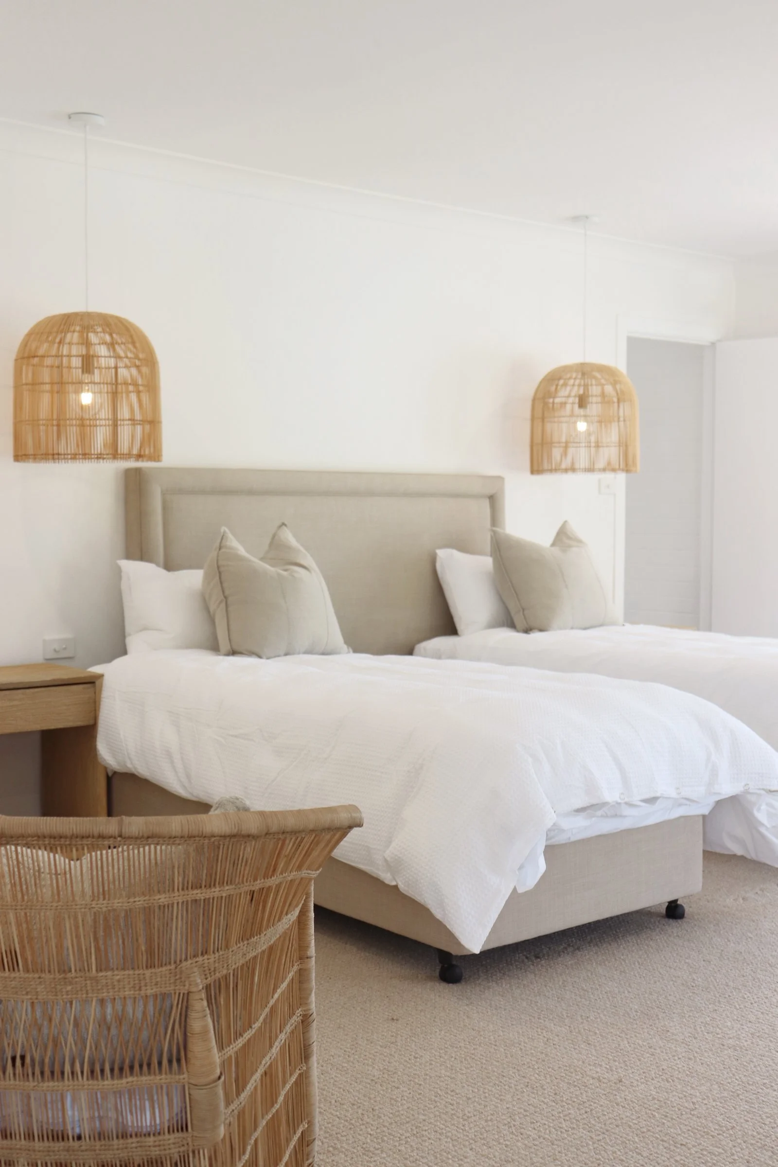 A hotel room with two double beds, beige headboards, white bedding, beige pillows, rattan pendant lights hanging from the ceiling, a wooden nightstand, and a wicker chair, with neutral color tones and minimal decor.