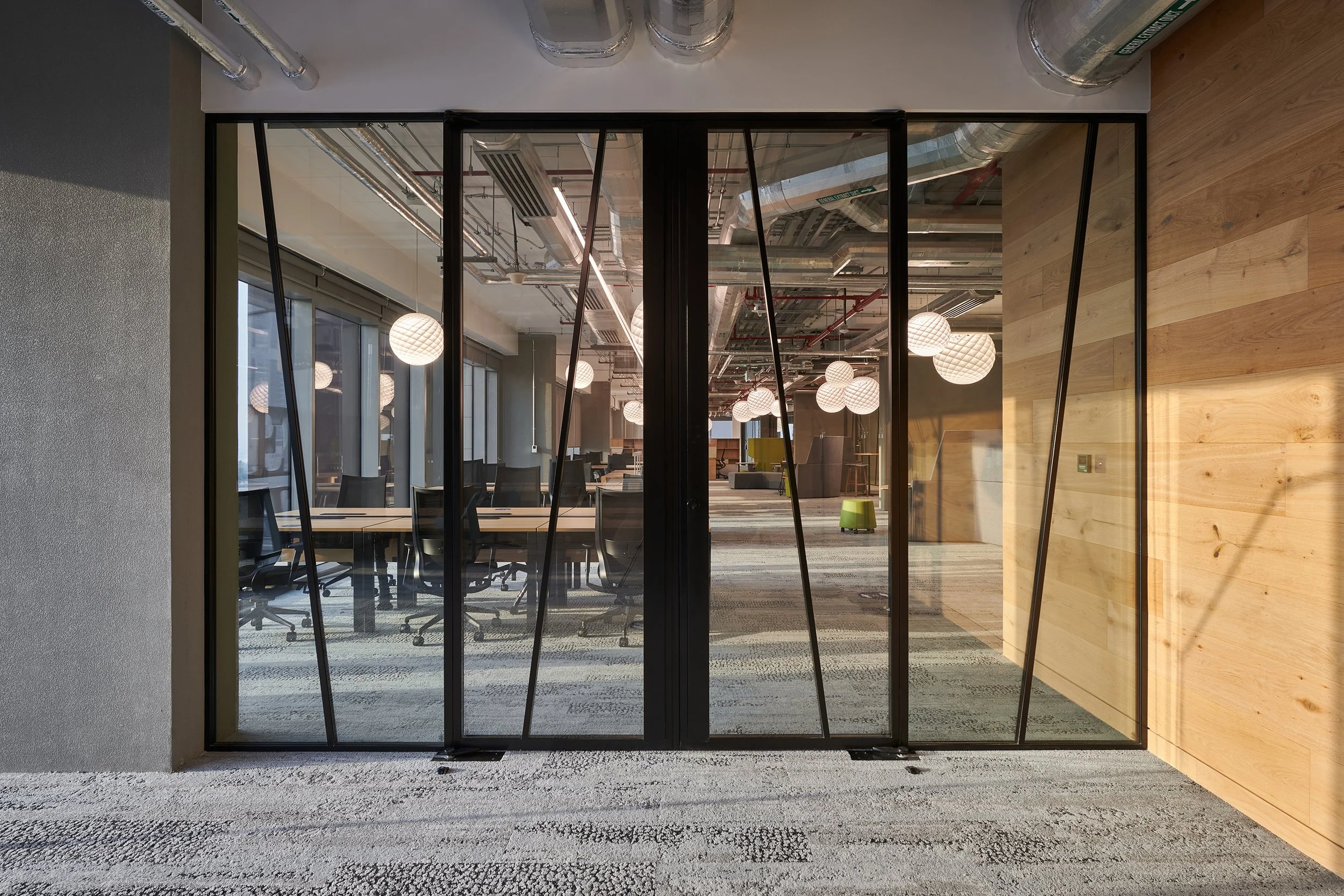 Modern office interior with glass walls, wooden accents, and open ceiling. There are hanging globe lights and conference tables with office chairs visible, creating a contemporary workspace atmosphere.