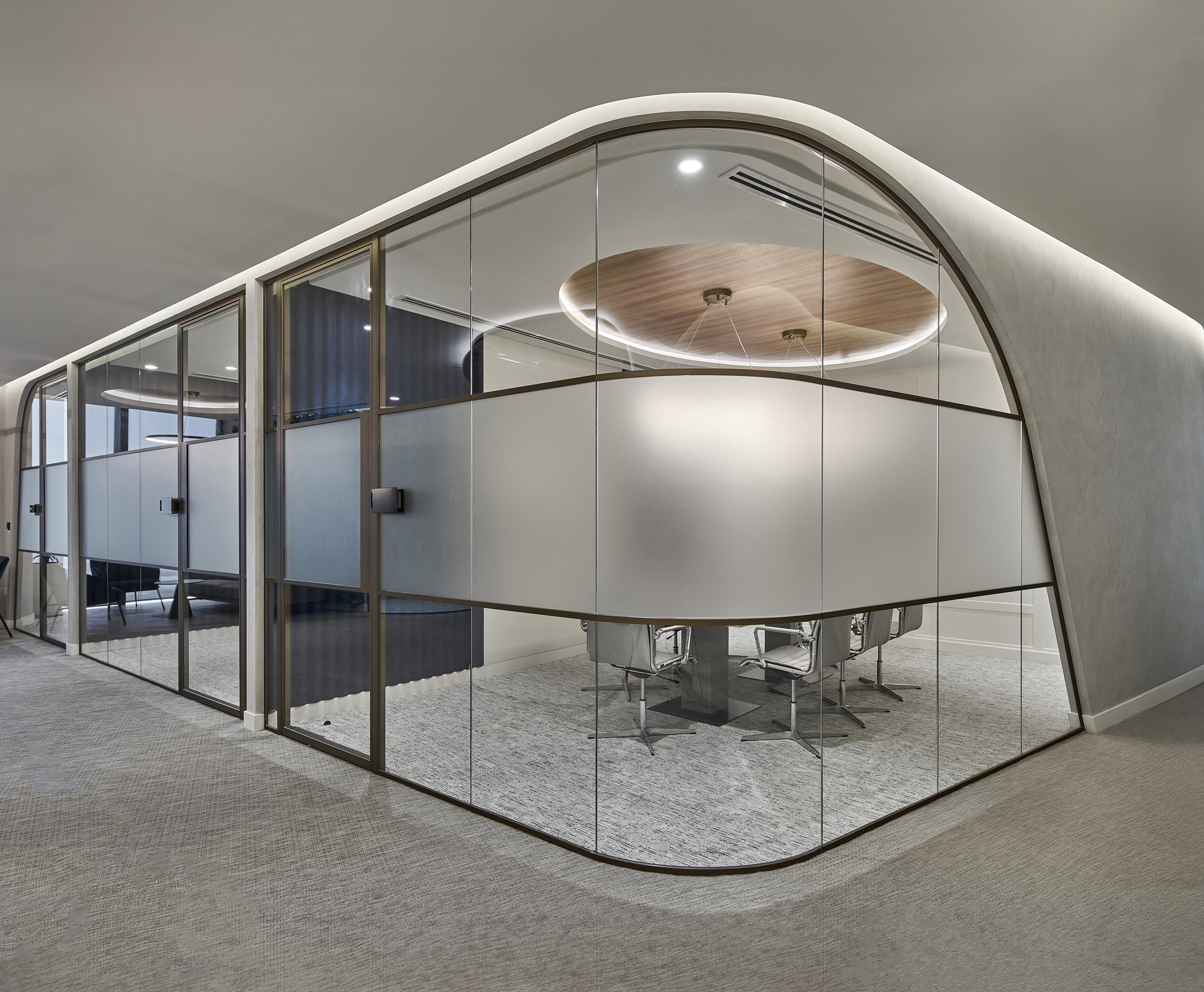 Modern office meeting room with curved glass walls and frosted panels, featuring a circular wooden ceiling design and sleek metal chairs around a table.