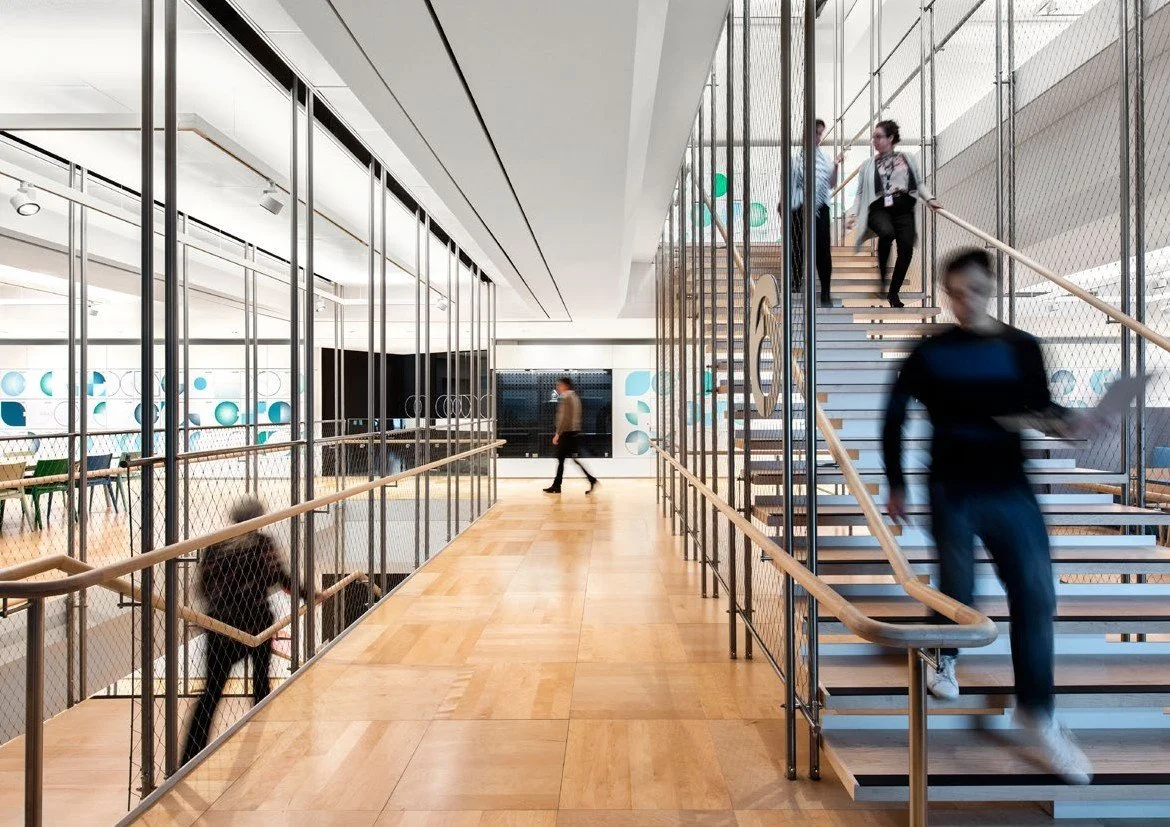 Modern office interior with people walking on stairs and corridor, featuring glass walls and wooden floors.