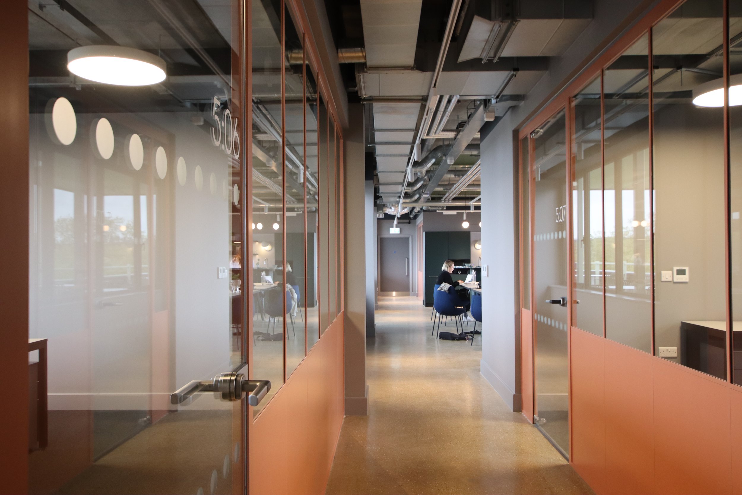 Modern office hallway with glass doors, numbered 506 and 507, leading to rooms with people working at desks.