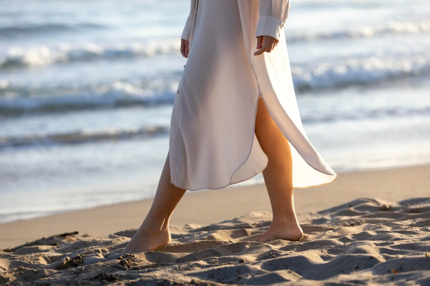 lower-portion-of-a-woman-with-bare-feet-walking-on-a-sandy-beach-with-waves-visible-in-the-background,-wearing-a-long-white-dress