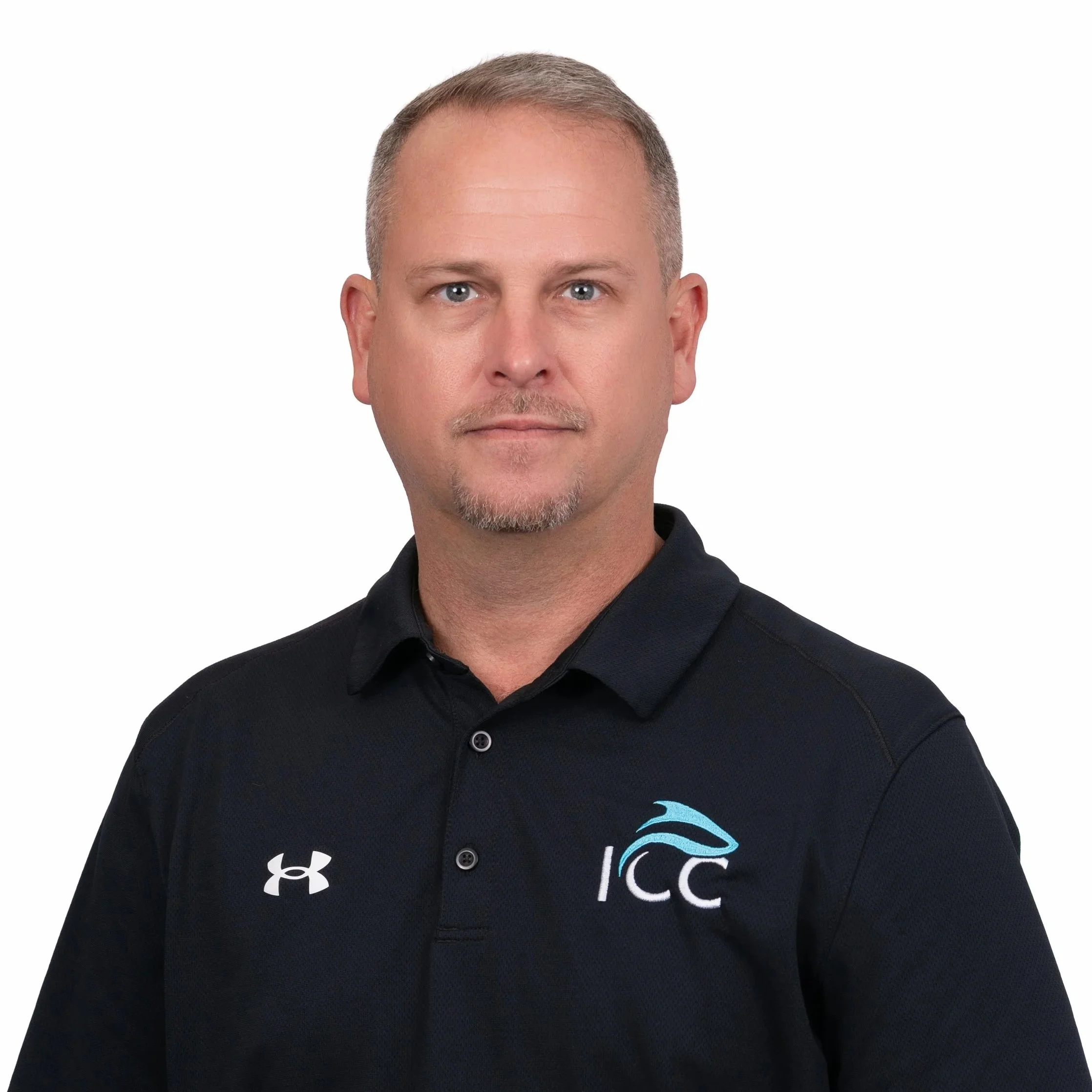 Portrait of a man with short blonde hair and blue eyes, wearing a black Under Armour polo shirt with an ICC logo on it, against a plain white background.