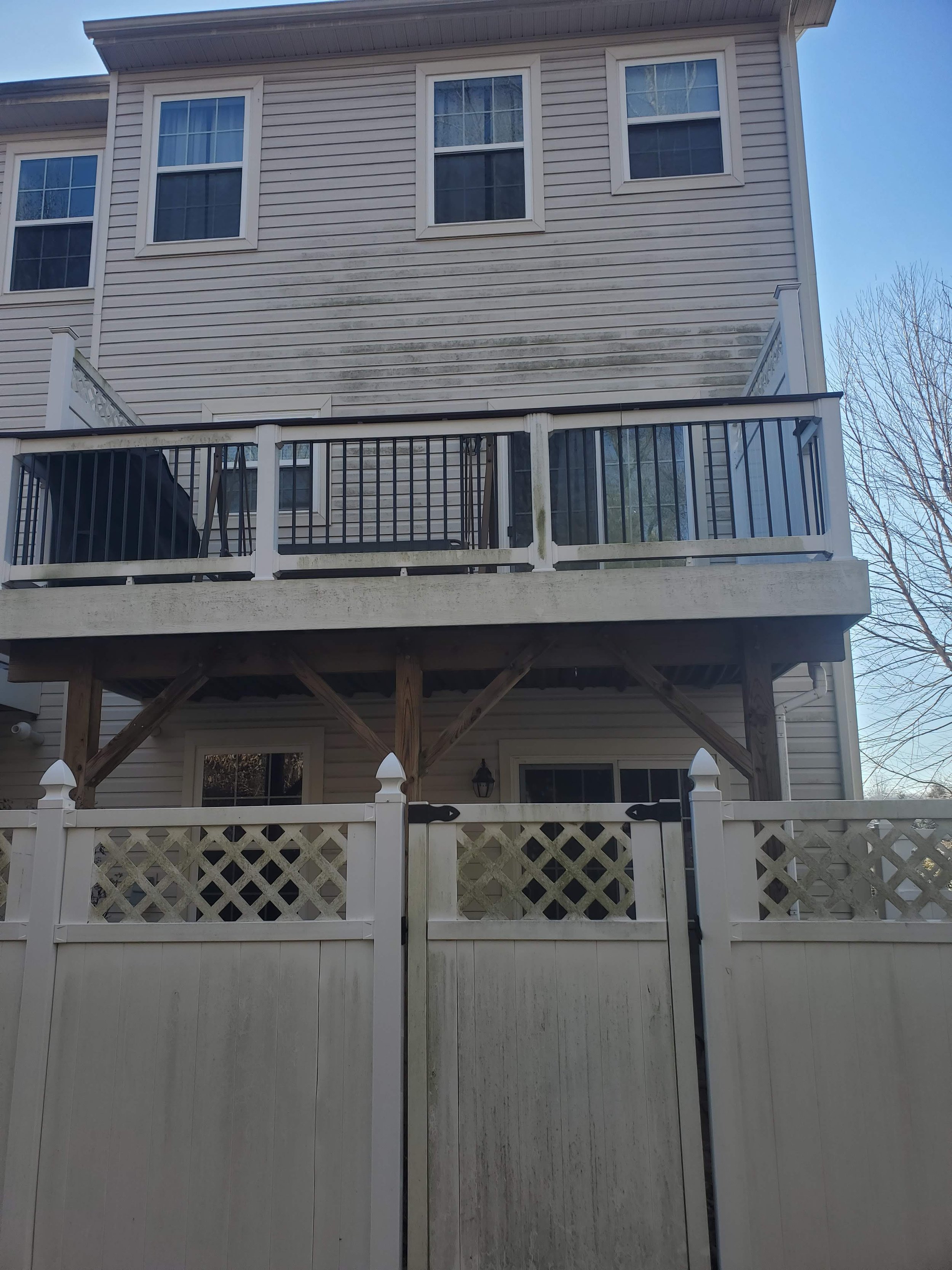 Two-story house with beige siding, three windows on the upper level, a deck with black and white railing, and a white lattice-topped fence in the foreground.