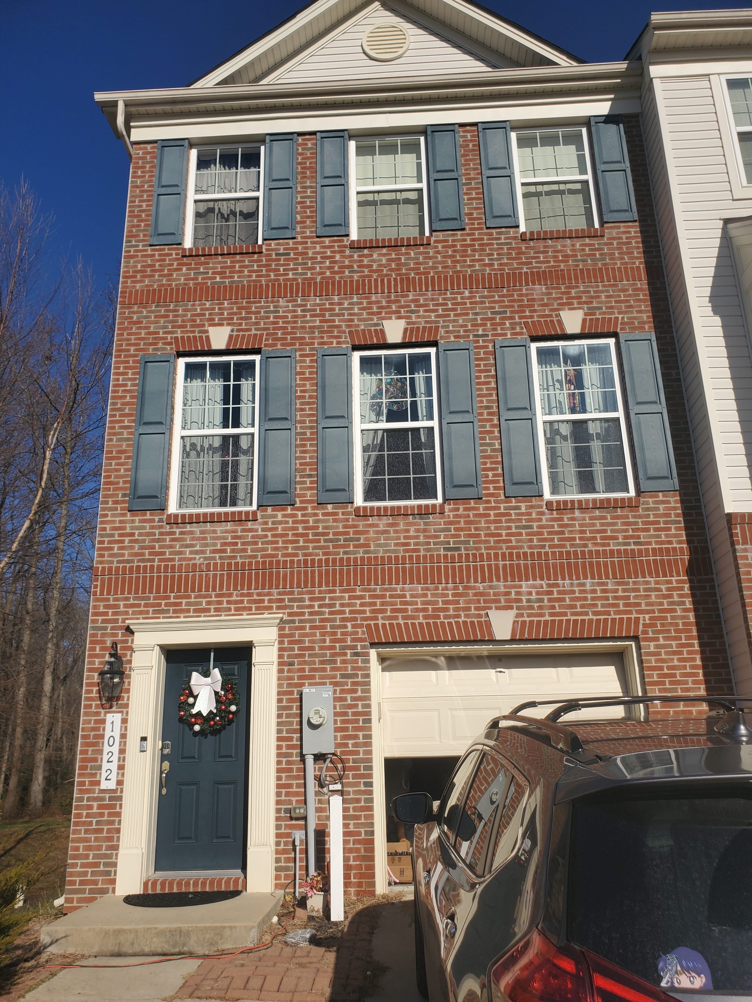 Brick townhouse with blue shutters, three stories, blue front door with holiday wreath, and attached garage. Gray SUV parked in driveway.