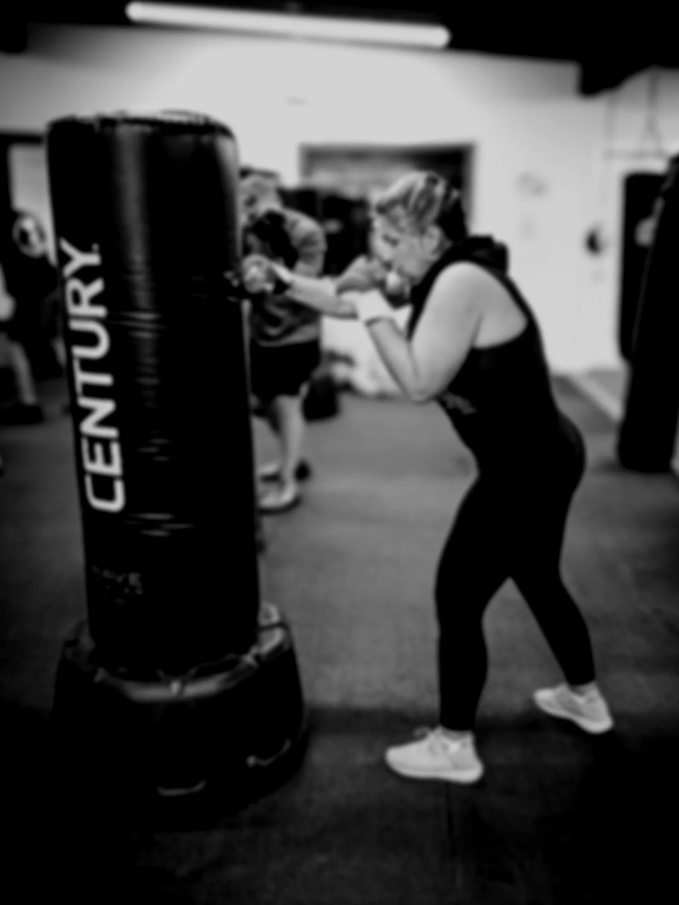 A woman practicing boxing with a heavy punching bag at a gym. She is wearing workout clothes and gloves, focusing on her training.