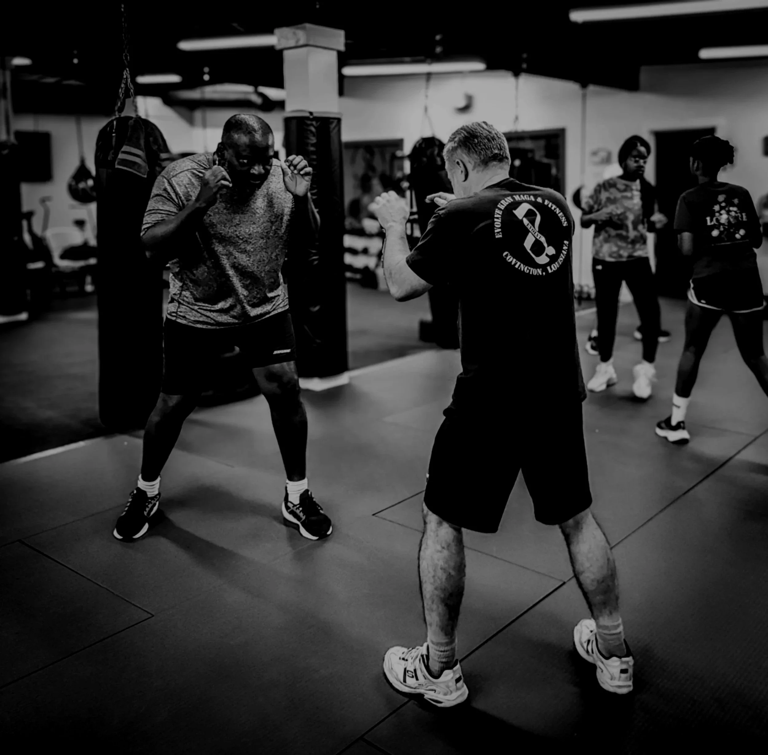 Two men sparring in a boxing gym, practicing defensive techniques while other individuals watch in the background.