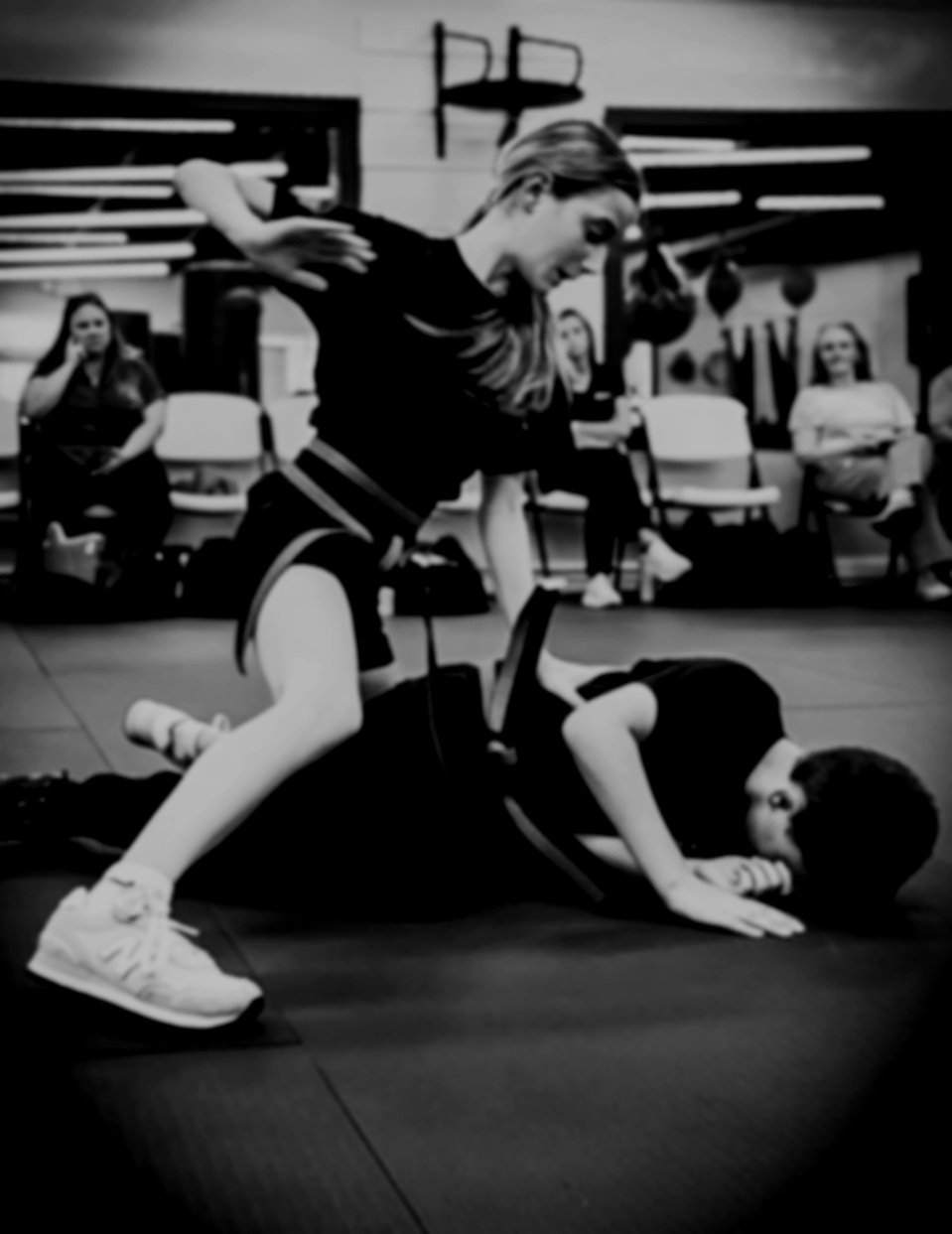 Two women practicing martial arts or self-defense techniques in a gym, with one woman on top of the other, in a classroom setting with certificates or awards in the background.