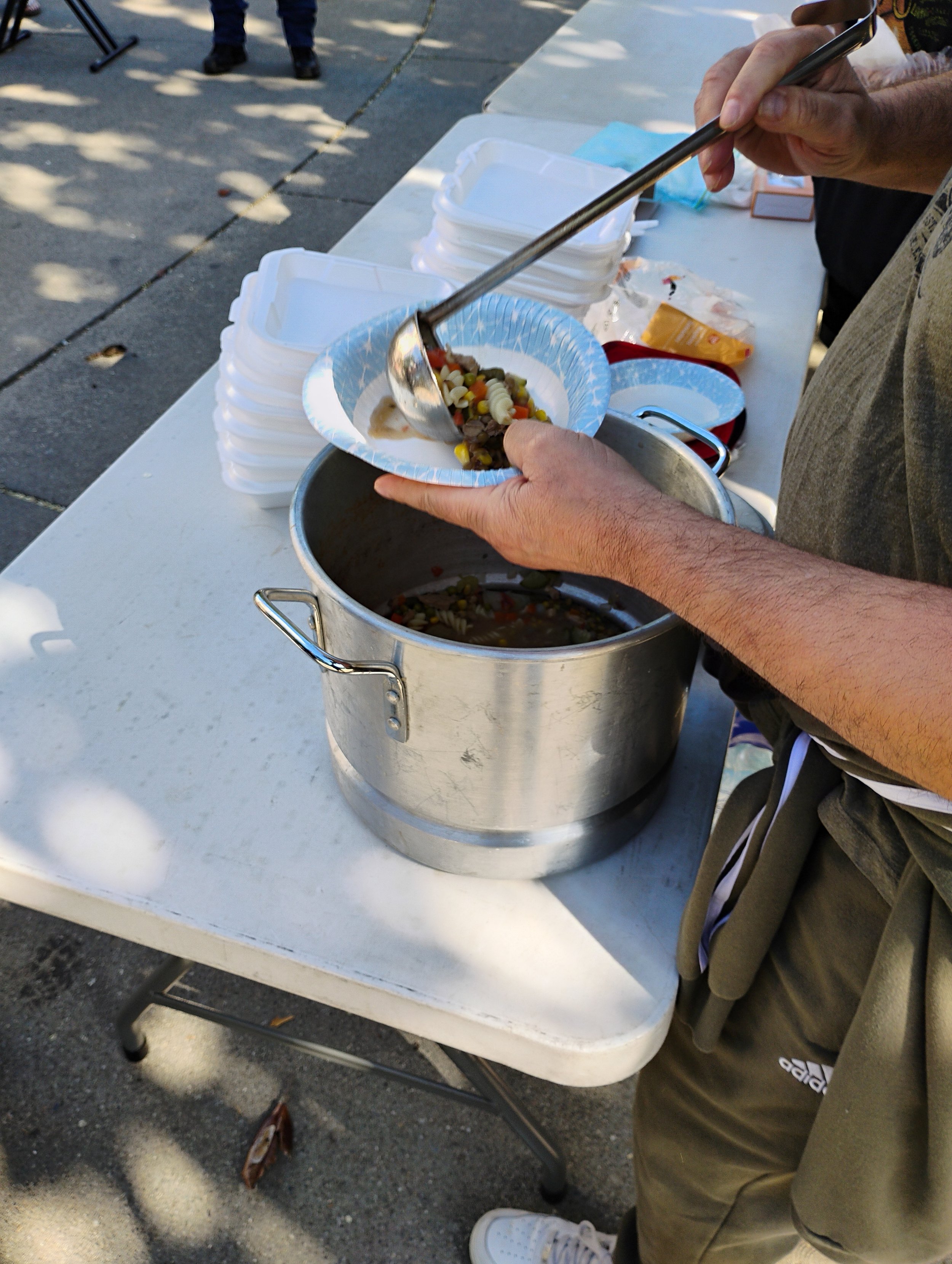 Person serving chili or stew from a large metal pot into a disposable plate at an outdoor event, with empty white disposable bowls stacked nearby on a table.