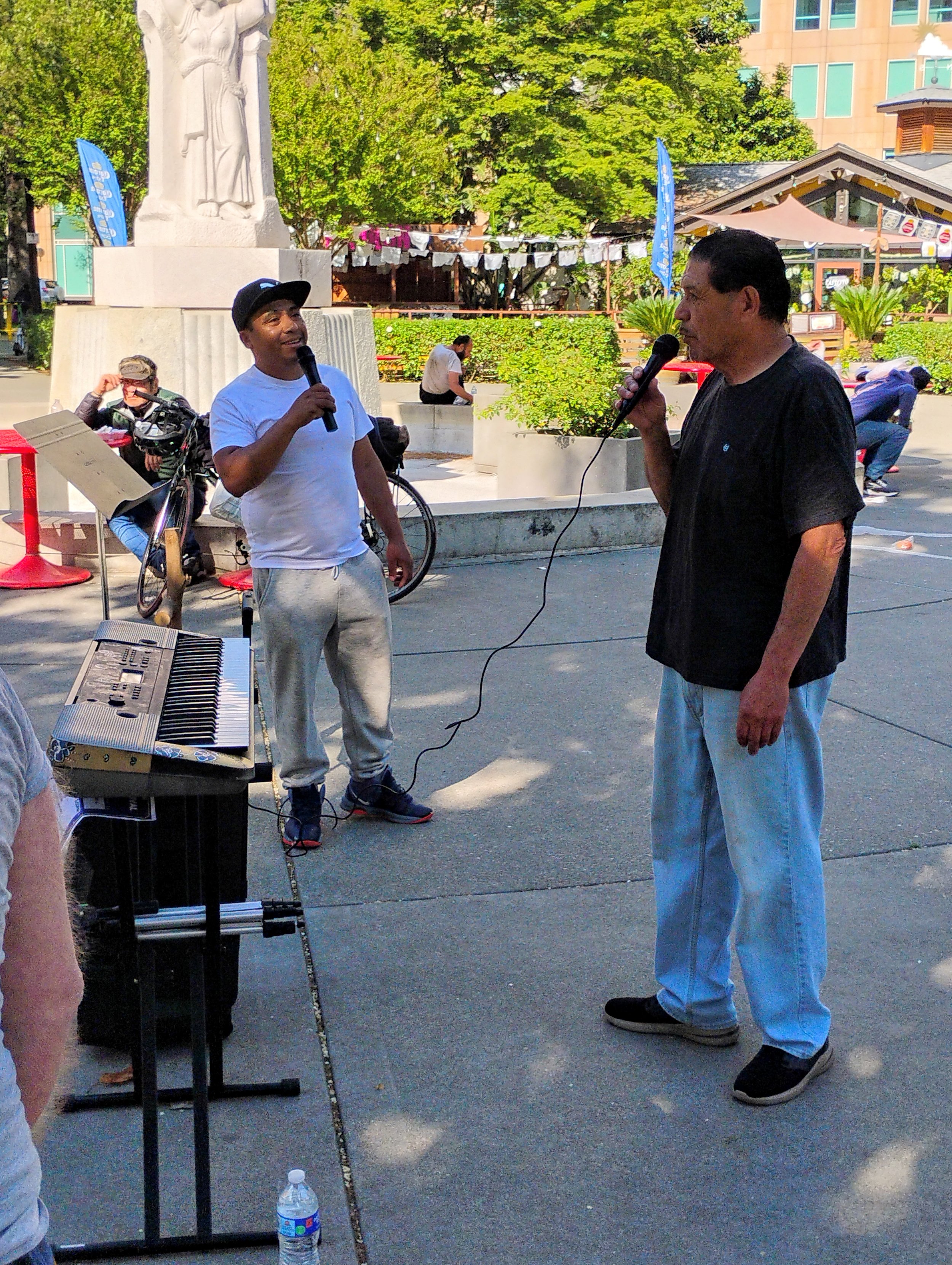 Two men are singing into microphones on a sidewalk in a park, with a keyboard and music stand nearby. One man is wearing a white t-shirt, gray sweatpants, and a black cap, while the other is dressed in a black t-shirt and light blue jeans. There are