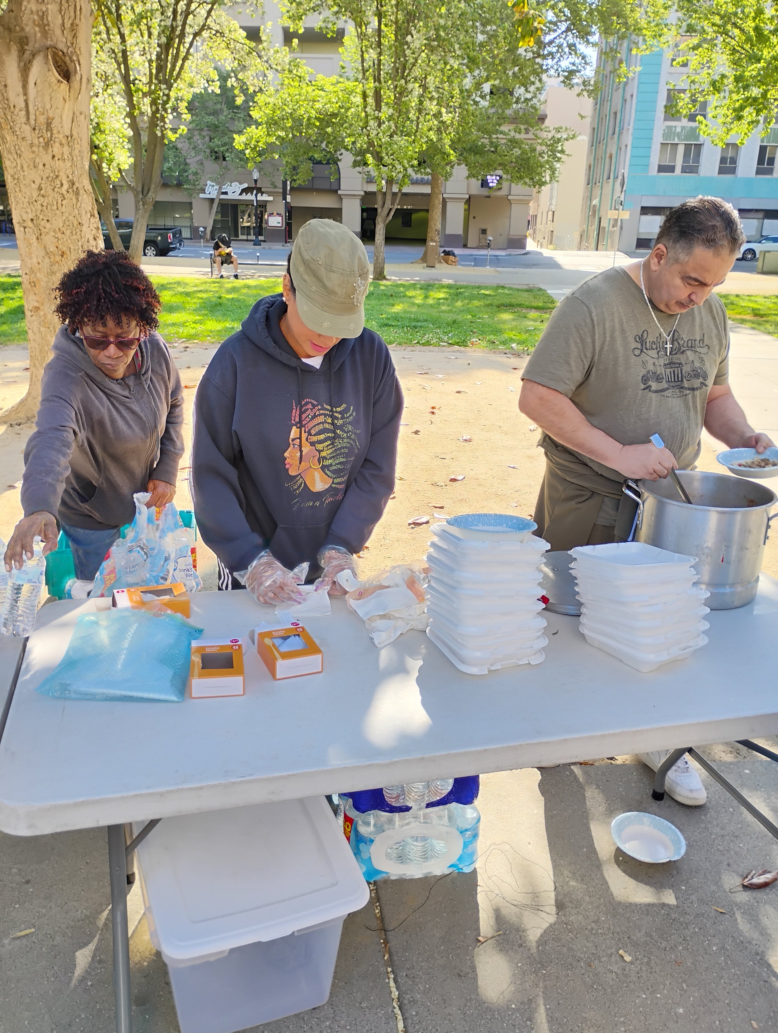 Three people standing at a table outdoors, serving food or drinks, with stacks of foam containers and bottled water on the table.