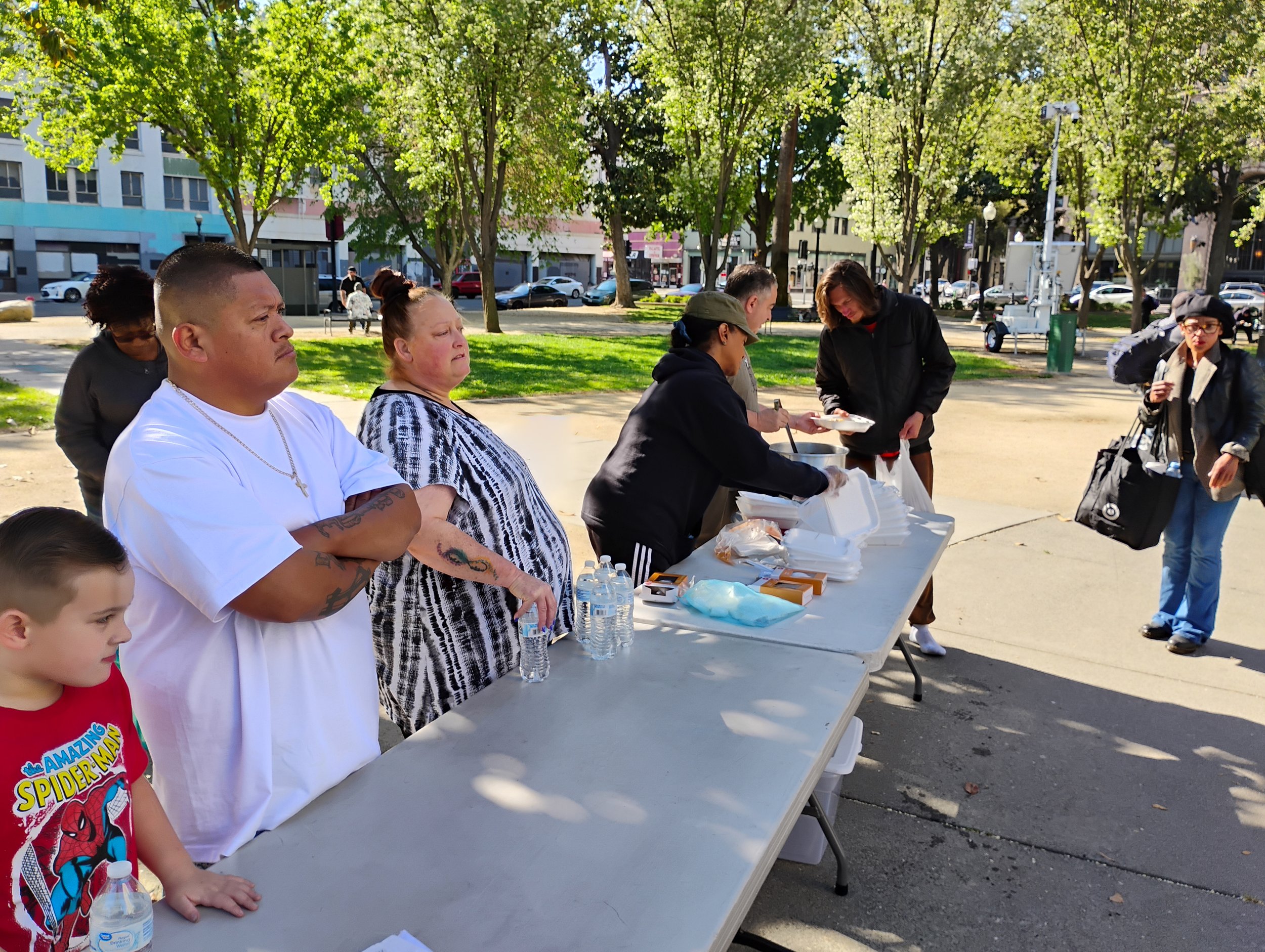 People lining up at an outdoor food distribution table in a park on a sunny day, with trees and buildings in the background.
