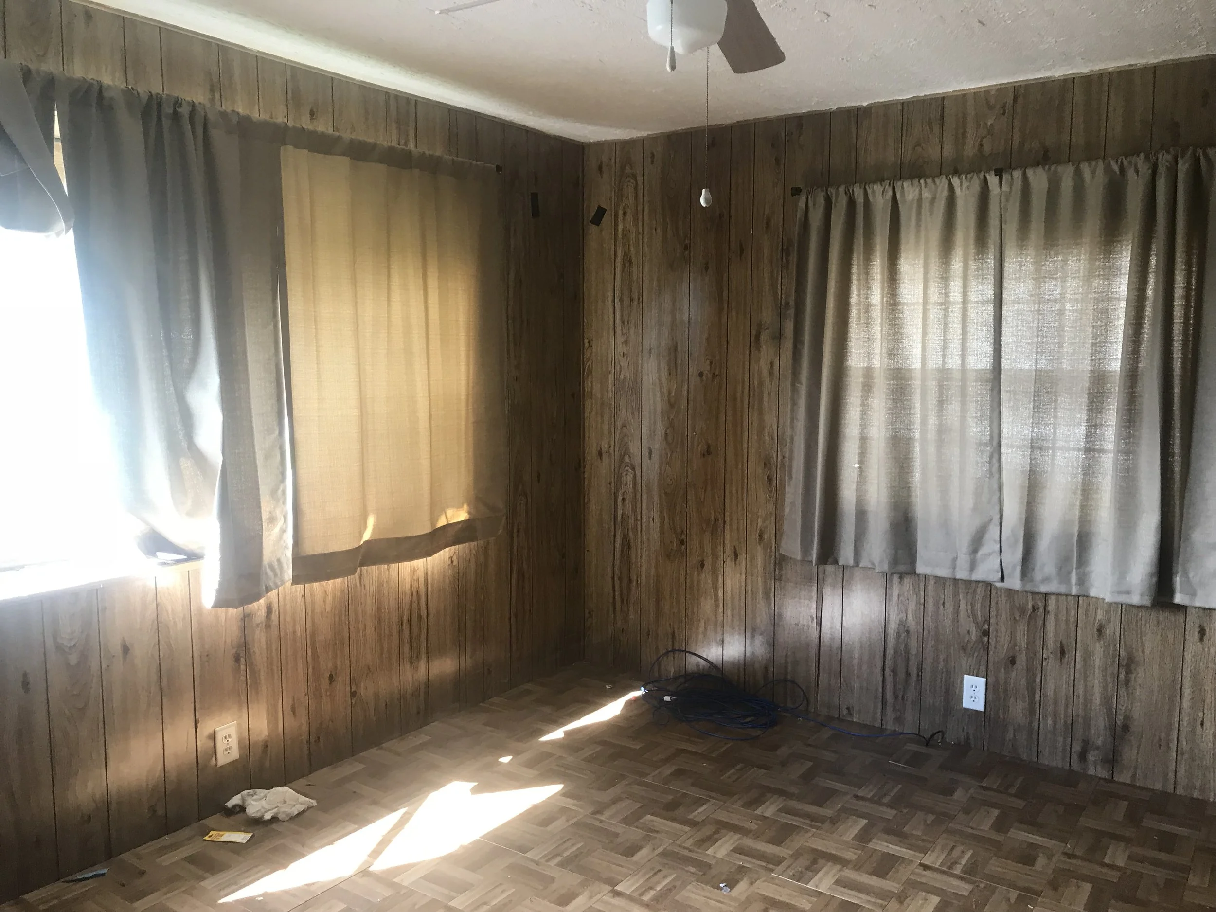 Empty room with wood paneling, parquet flooring, and beige curtains. Picture is of a bedroom before remodel was complete. 