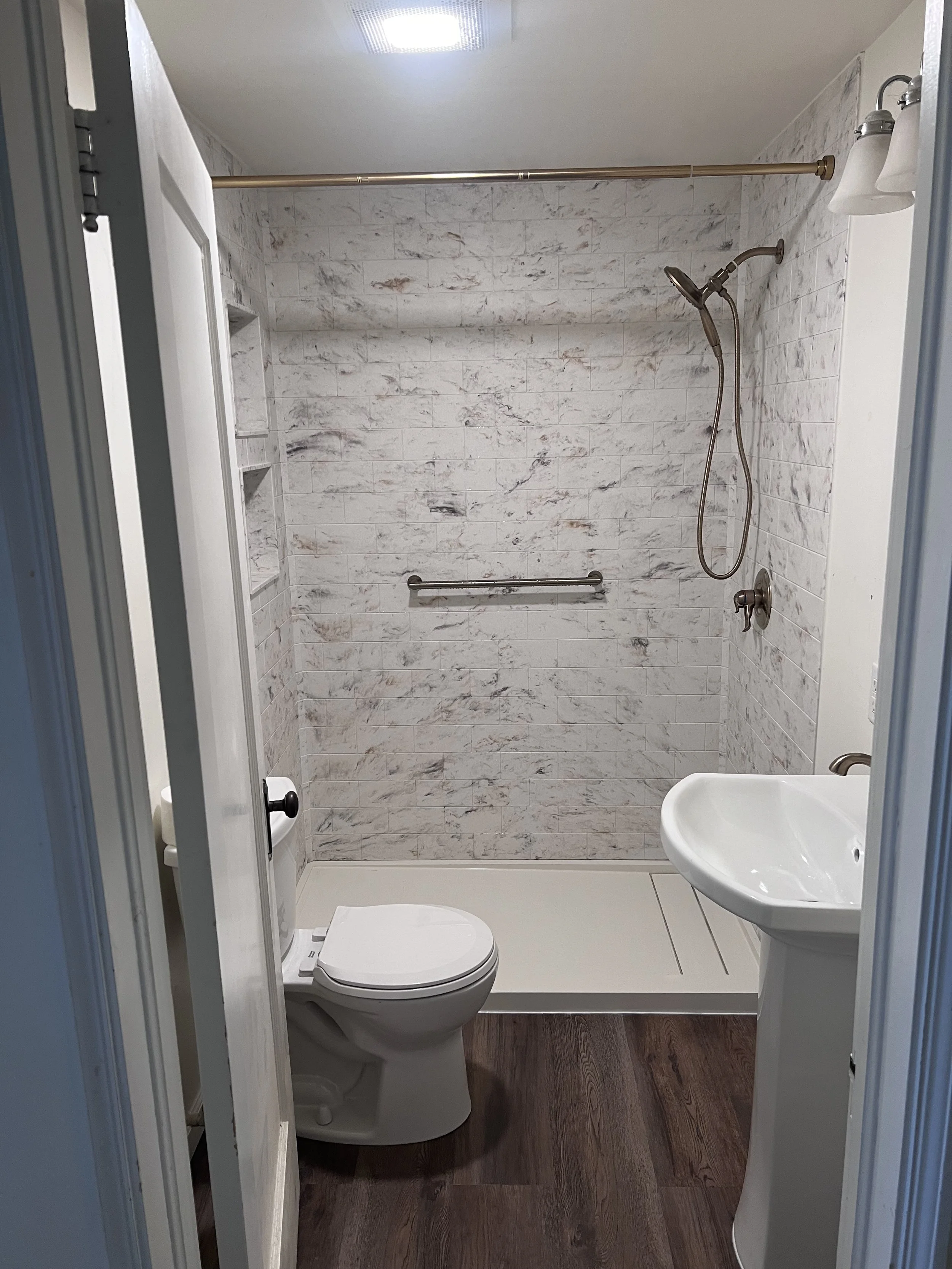 Modern bathroom with a white toilet, pedestal sink, and tiled walk-in shower. Features dark wood flooring, a handheld showerhead, a silver grab bar, and white walls with a marble pattern. The room is lit by a ceiling light and a wall-mounted fixture.