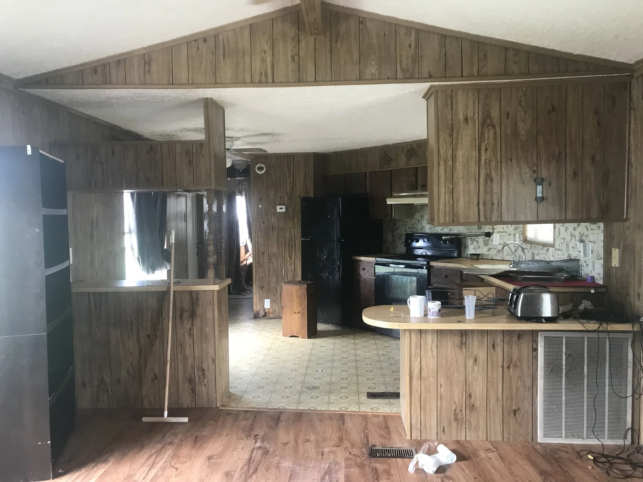 Interior of an older kitchen with wood paneling, laminate floors, and cluttered countertops. Appliances include a refrigerator, stove, and toaster. Picture is of a kitchen before kitchen remodel was complete. 