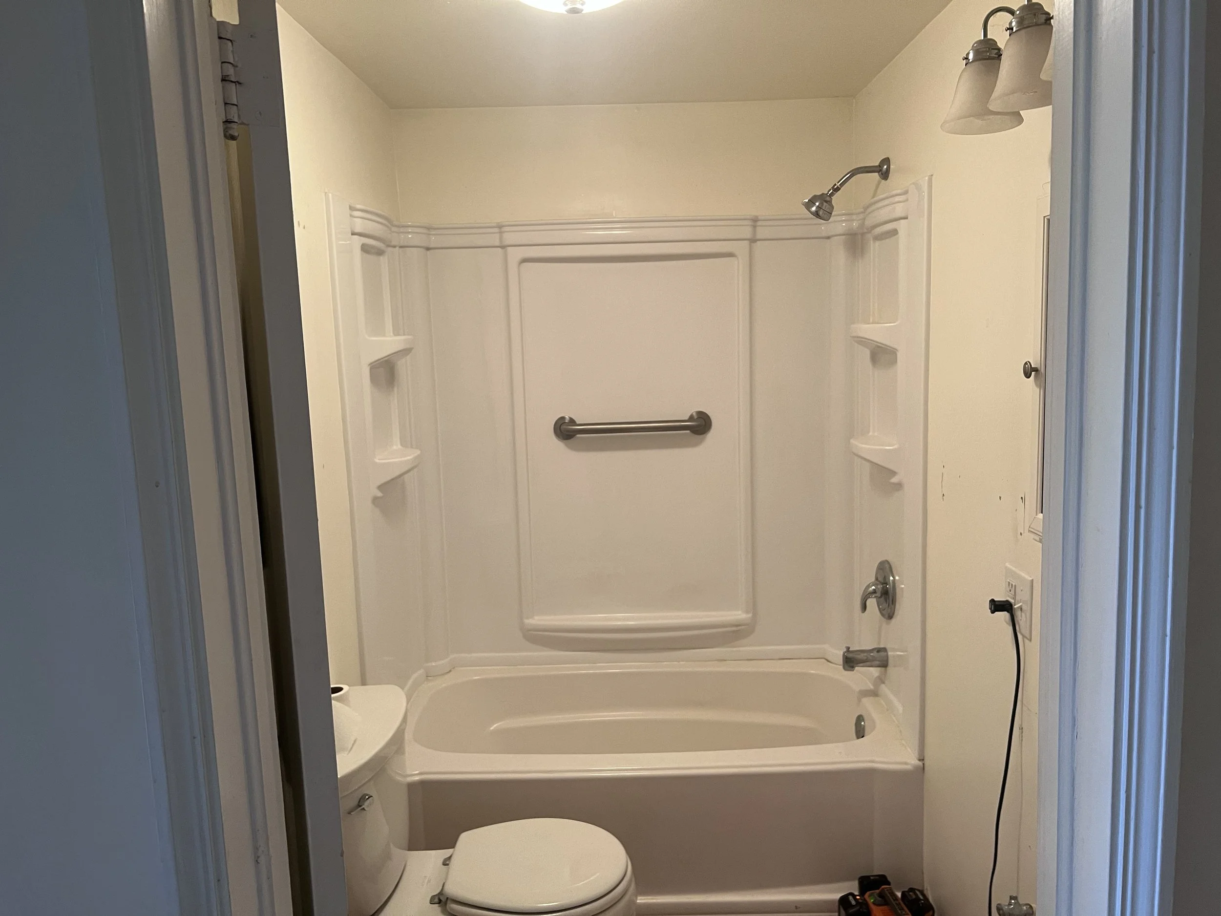 Small bathroom with a white bathtub-shower combo, toilet, and light fixture. It includes wall shelves, a grab bar, and a showerhead over the tub. The room has cream-colored walls and a door slightly open.