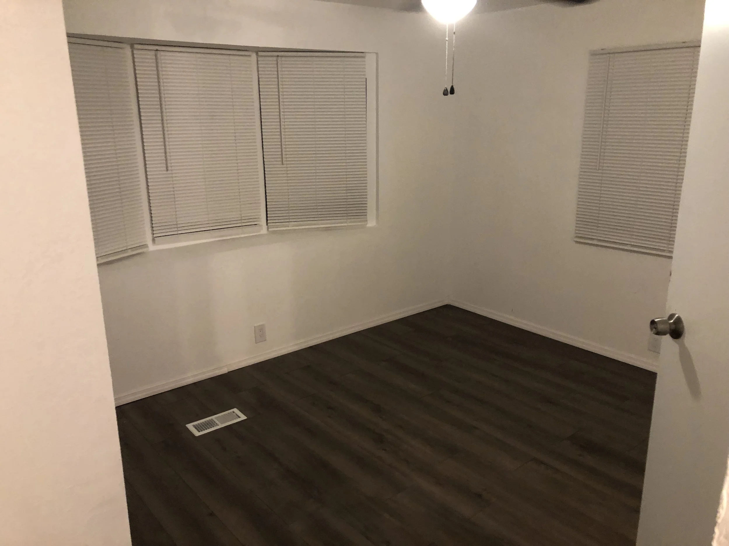 Empty room with dark wood flooring, white walls, and three windows with closed blinds.
