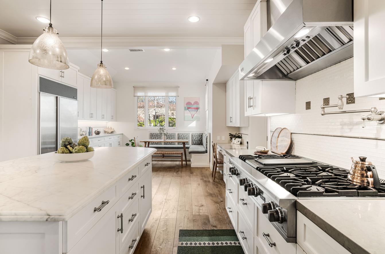 Bright kitchen with white cabinets, marble countertop island, stainless steel stove, and wooden dining nook with window and artwork.