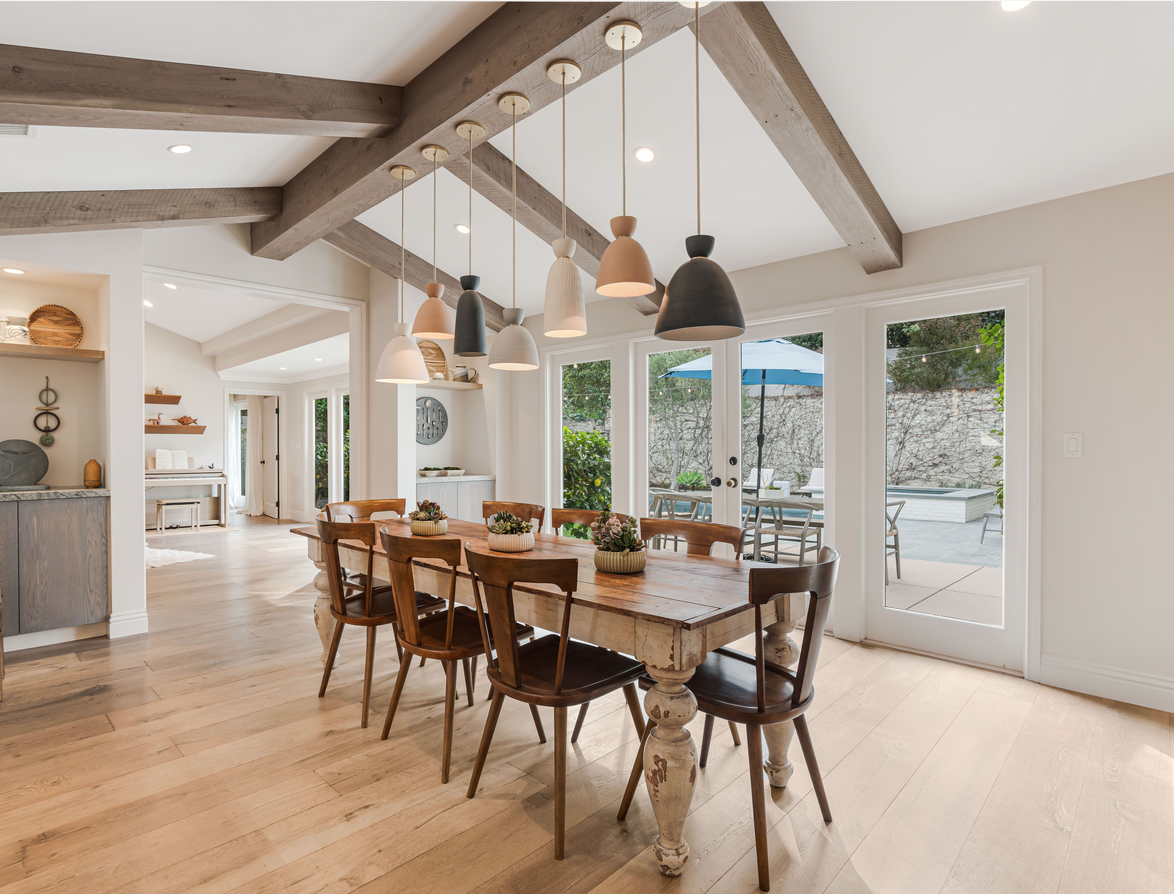 Dining room with wooden table and six chairs, hanging light fixtures, and large glass doors leading to an outdoor patio with seating and umbrella.