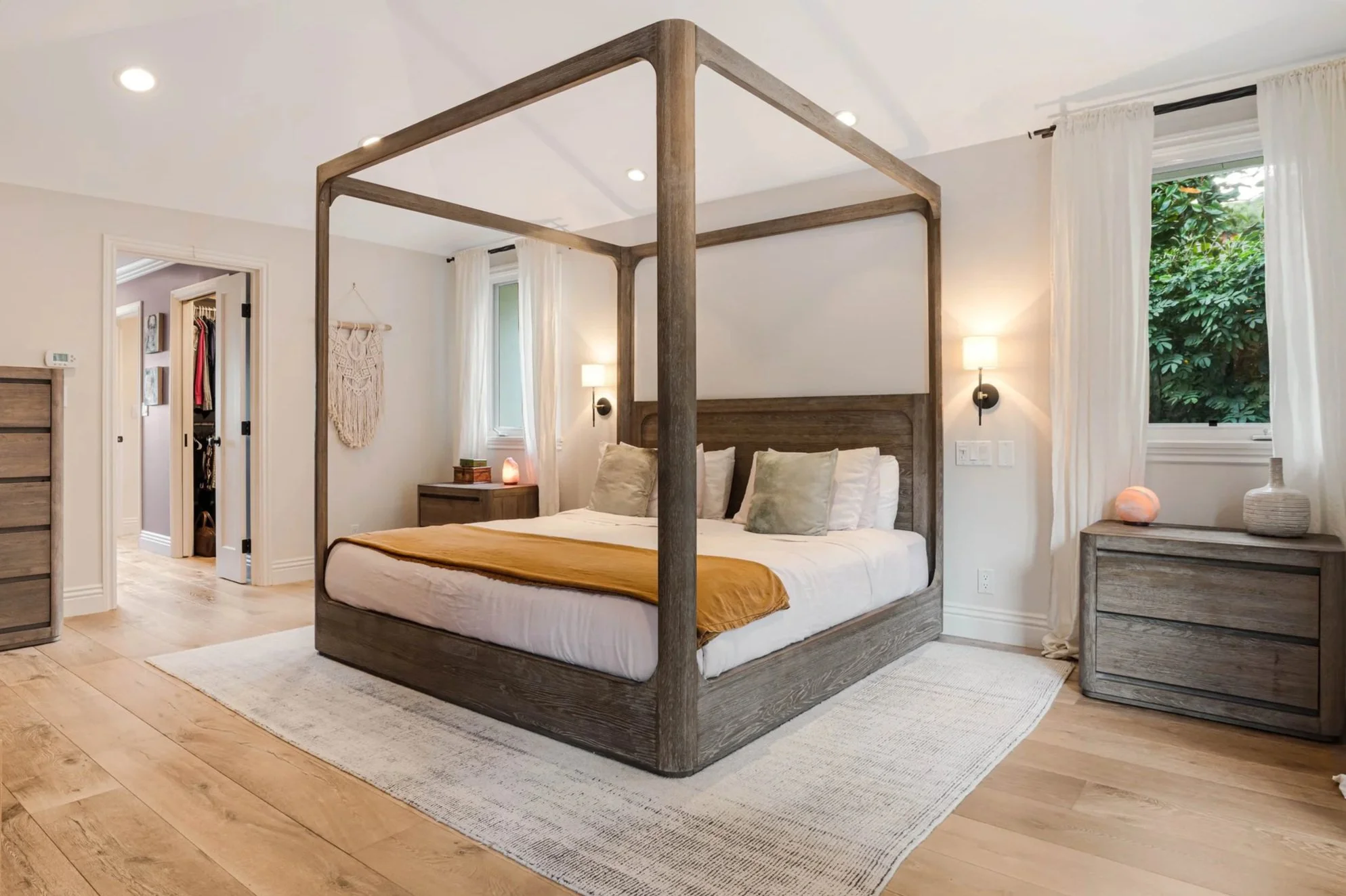 A bedroom with a wooden four-poster bed, cream-colored bedding, and decorative pillows. There are two nightstands with lamps and a large window with white curtains. The floor is wood, and there is a light-colored rug.