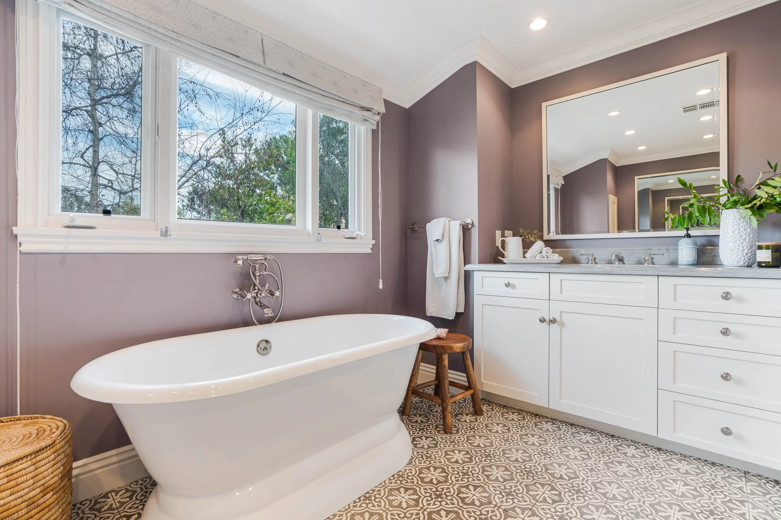 A bathroom with a large window showing trees outside, a white bathtub, a wooden stool, a white vanity with a large mirror, a potted plant, and a towel hanging on a metal rod.