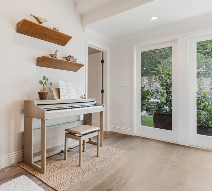 A white digital piano with a matching bench, a plant in a pot, and sheet music on the music stand are in a bright room. Two floating wooden shelves with decorative animal figurines are on the wall above the piano. Large glass doors reveal a view of greenery outside.