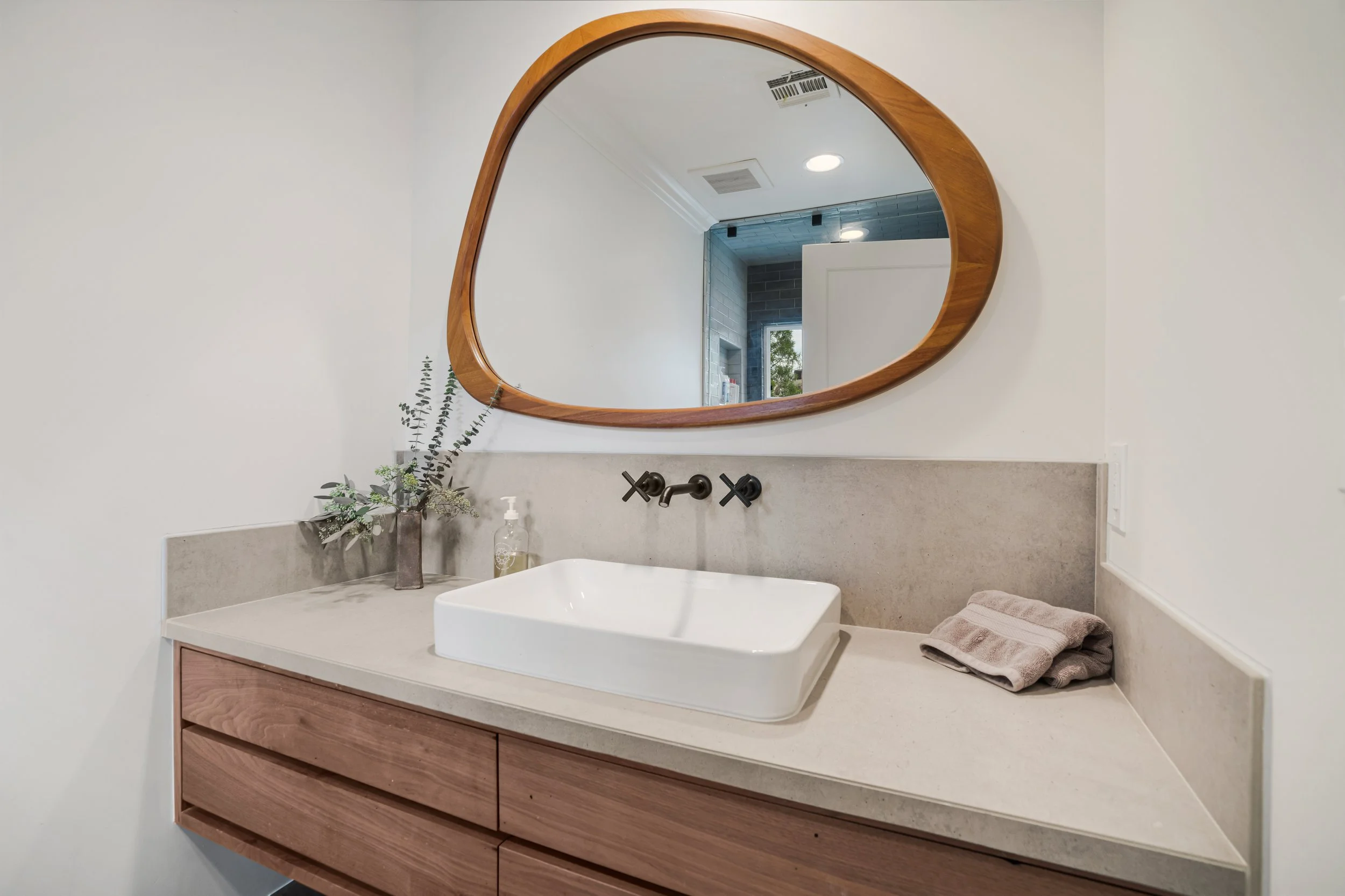 Bathroom vanity with a white vessel sink, a wooden cabinet, a mirror with a wooden frame, a faucet, a small vase with greenery, a soap dispenser, and a towel on the right side.