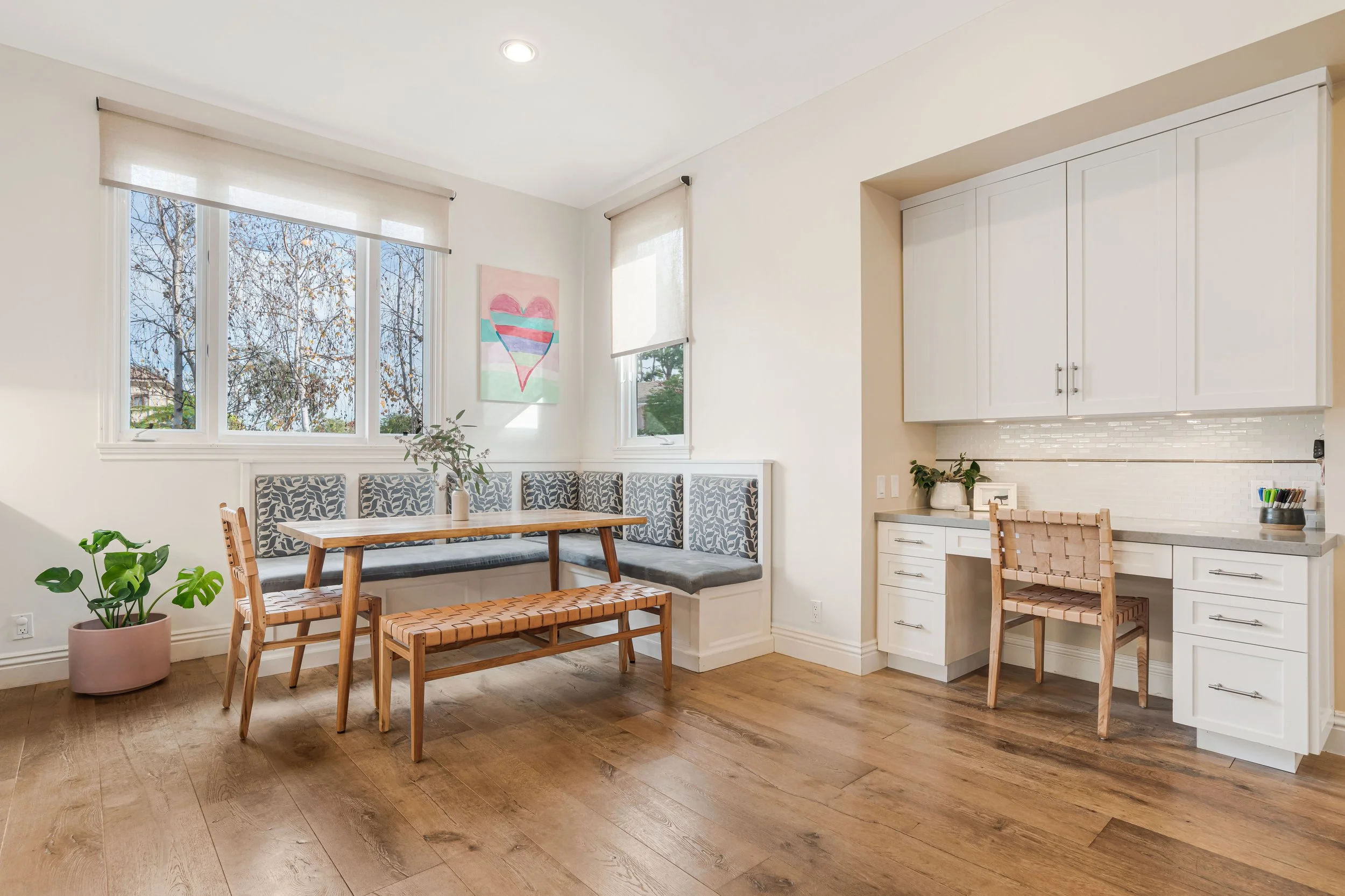 Bright kitchen and dining area with large windows, white cabinets, wooden dining table, woven chairs, indoor plants, and a colorful heart artwork on the wall.