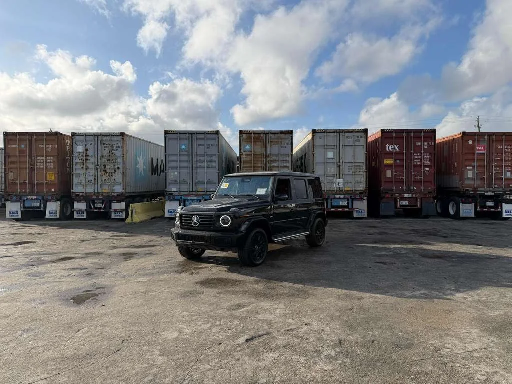 Black Mercedes-Benz G-Class SUV parked in front of a row of stacked cargo containers on a large, paved lot with a partly cloudy sky.
