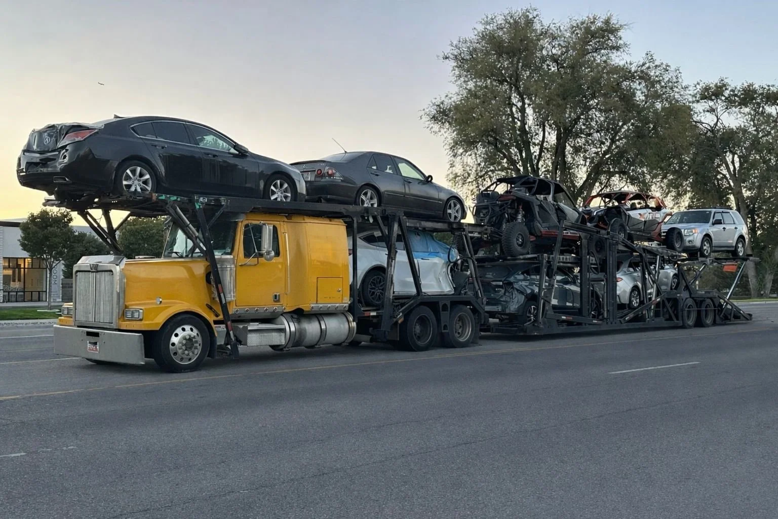 A large yellow car carrier truck transporting multiple black, gray, and white cars on a road with trees and buildings in the background.