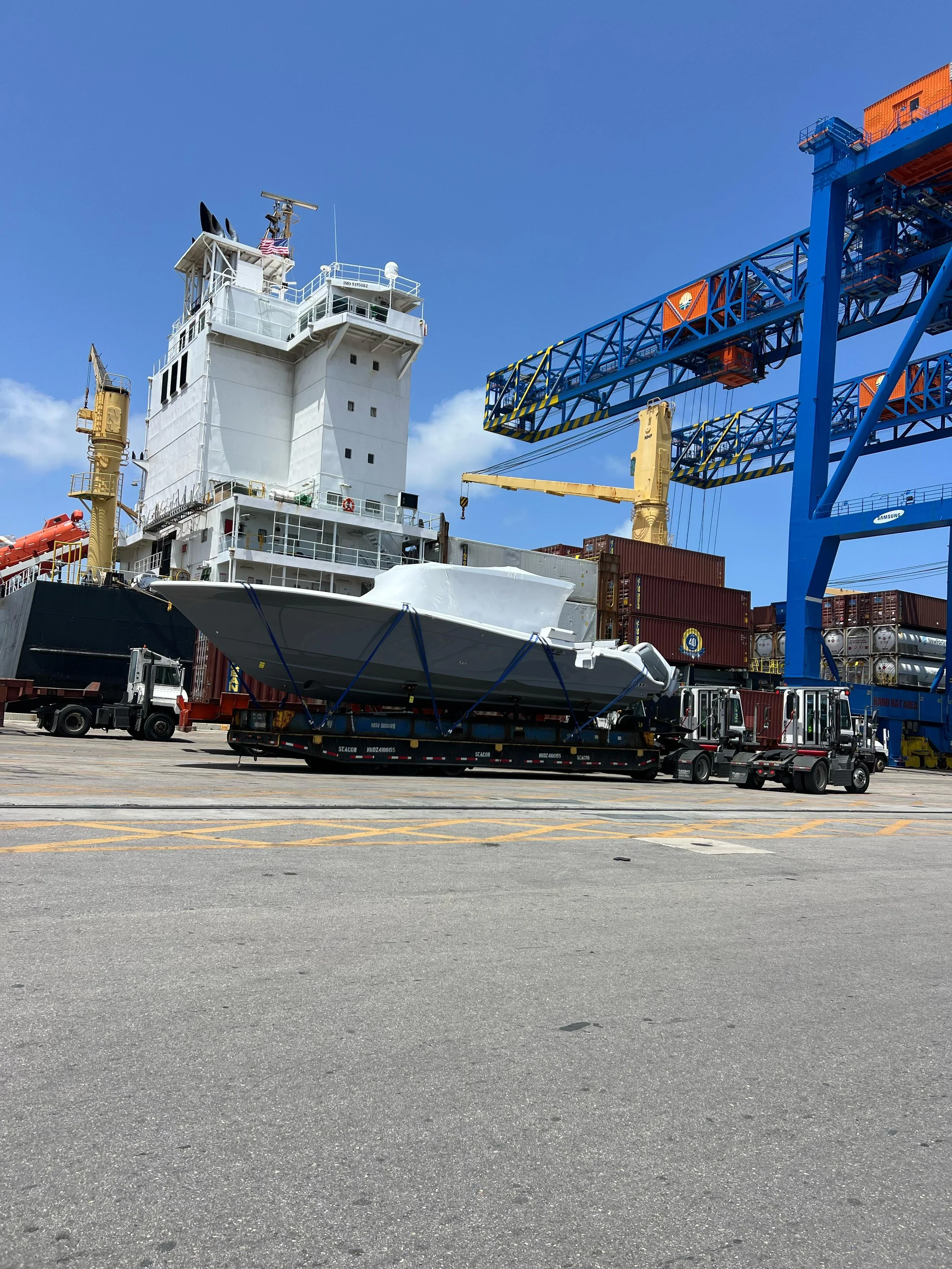 A ship being loaded at a port with a large white structure on a flatbed, accompanied by multiple cranes and shipping containers under a clear blue sky.