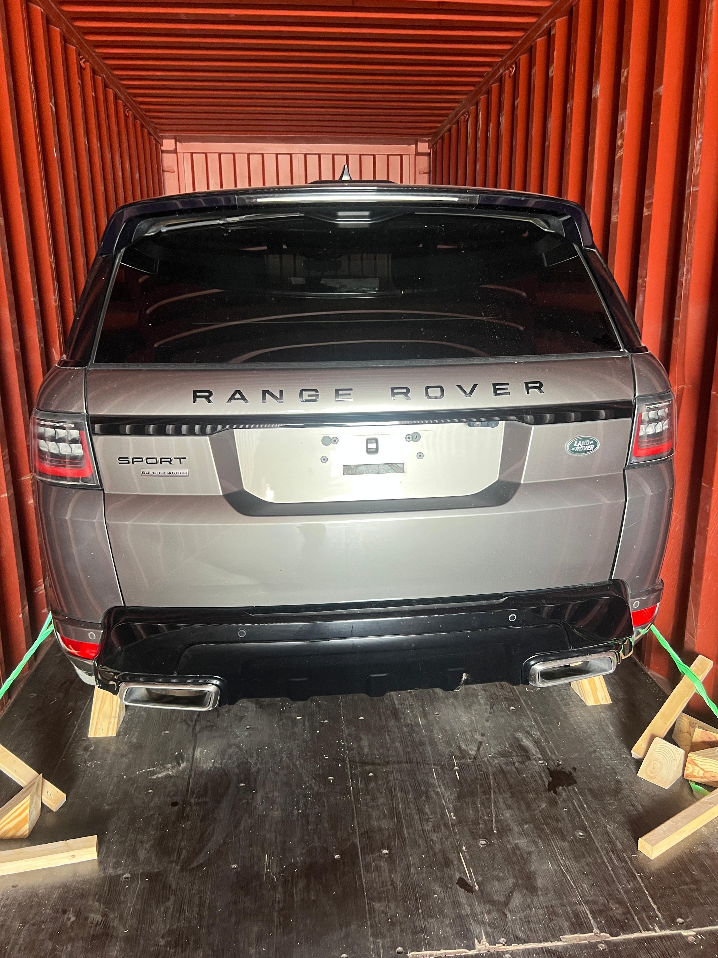 Rear view of a silver Land Rover Range Rover Sport parked inside a red shipping container, secured with wooden blocks and green straps.