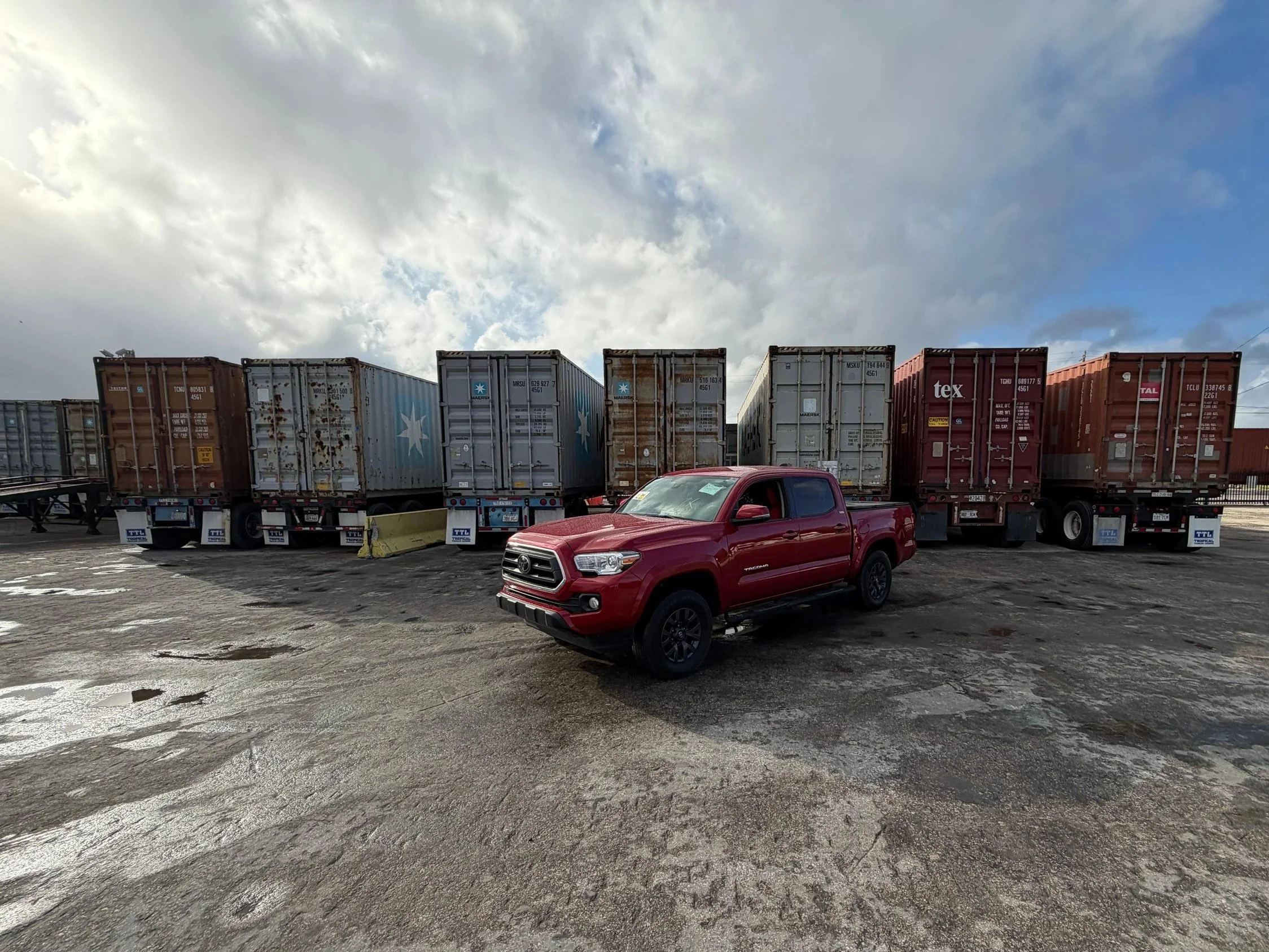 A red pickup truck parked in front of a row of shipping containers on a concrete lot under a partly cloudy sky.