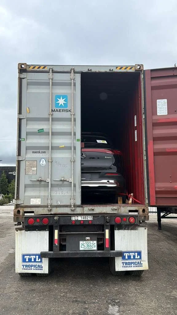 A car inside a shipping container on a truck, secured with a red strap.