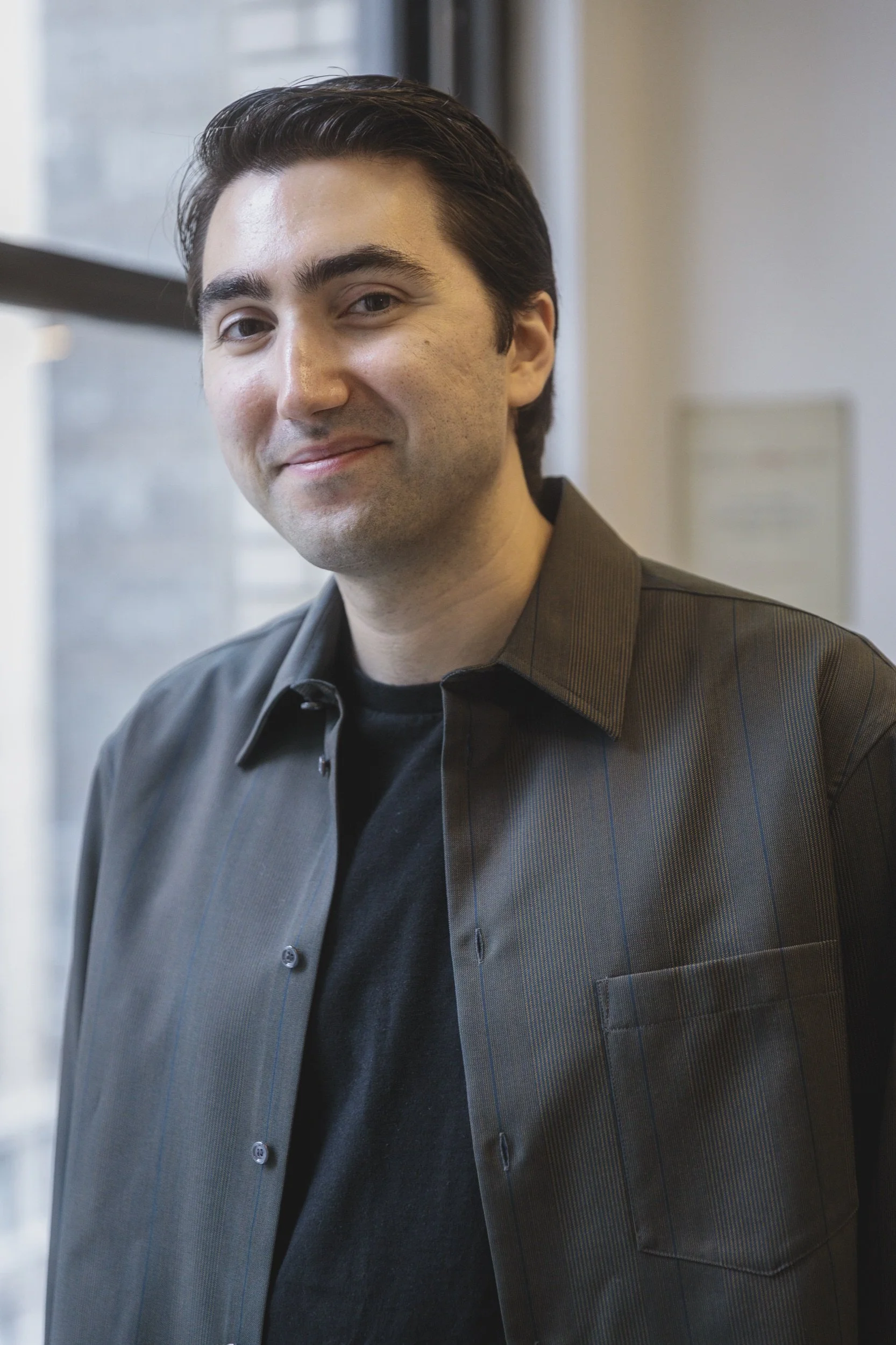 Alex Wakim with dark hair and light skin, wearing a dark gray button-up shirt over a black t-shirt, smiling slightly while standing indoors by a window.