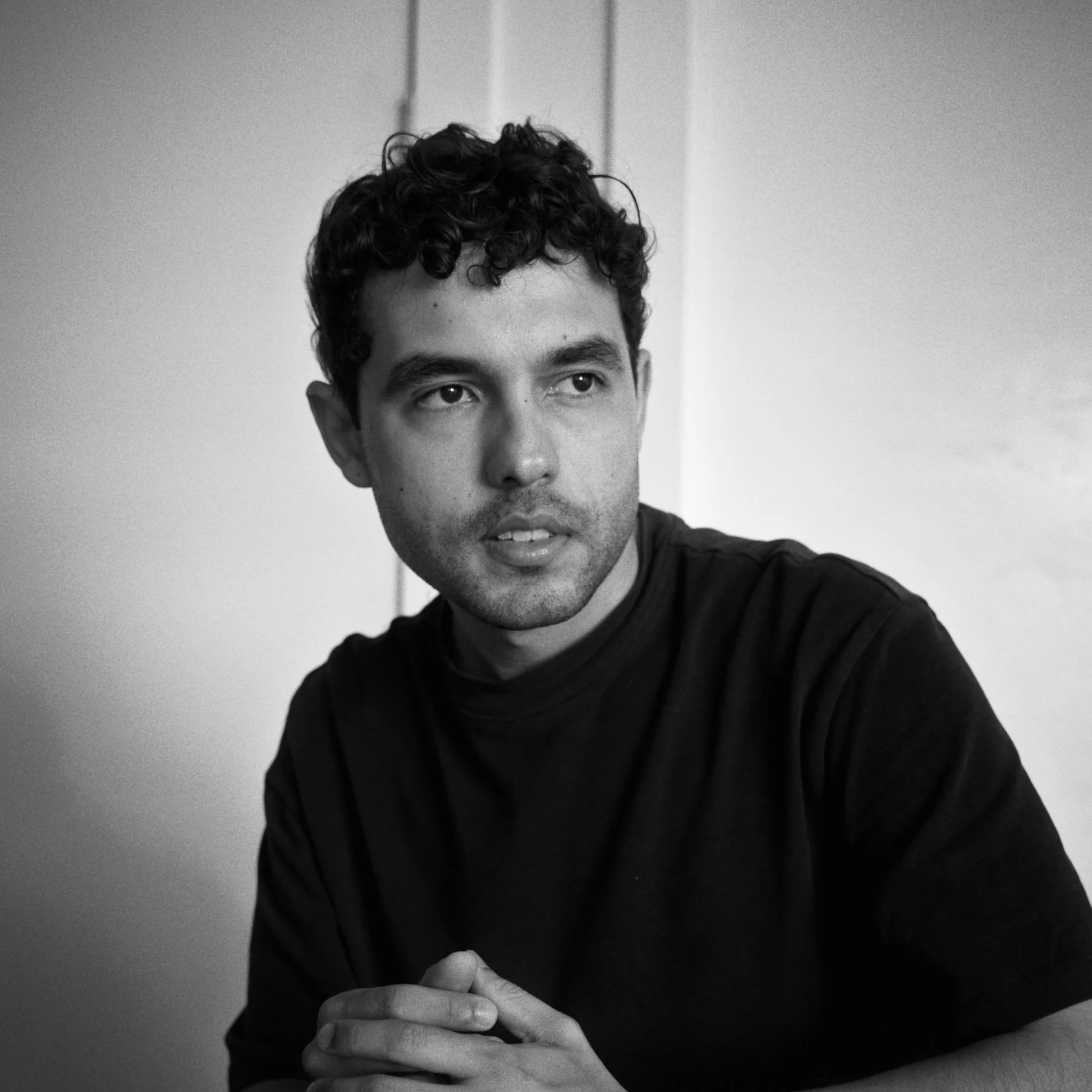 A black and white portrait of Juan Dussan, wearing a dark shirt, resting his chin on his folded hands, looking thoughtfully to the side.