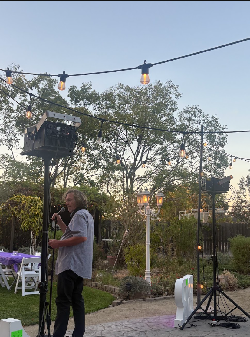 A person with long, curly hair holding a microphone and standing near sound equipment outdoors during twilight, surrounded by string lights and trees.