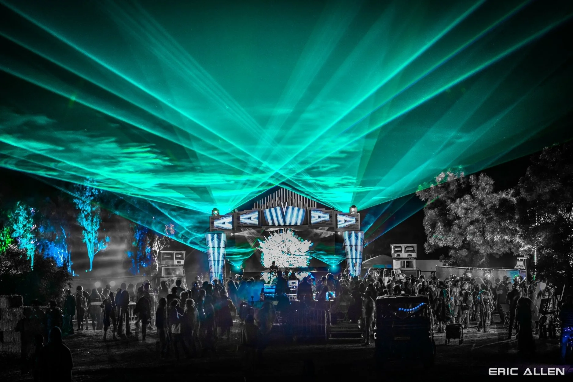 Nighttime outdoor concert with laser light show featuring blue and green beams overhead. Audience gathered in front of stage with trees illuminated by lights in the background.