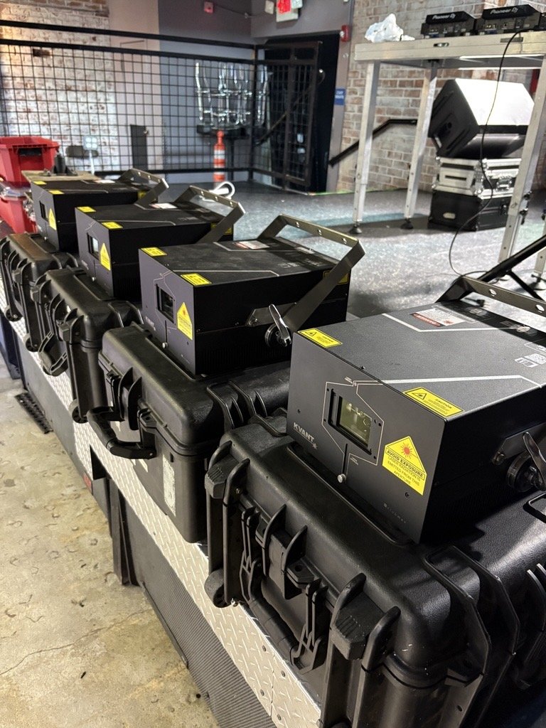 Row of laser projectors on a table at a nightclub or event venue, with equipment and a brick wall in the background.